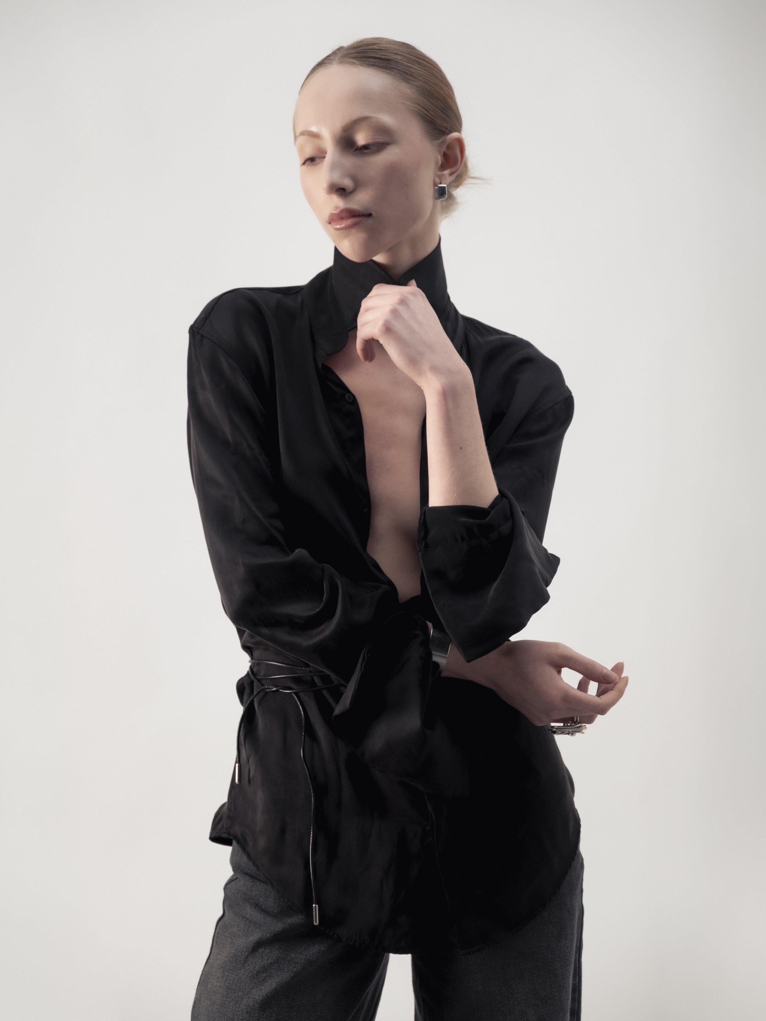 Une femme avec des cheveux attachés en chignon, portant une chemise noire en satin, une jupe noire, et des boucles d'oreilles carrées, posant contre un fond blanc.