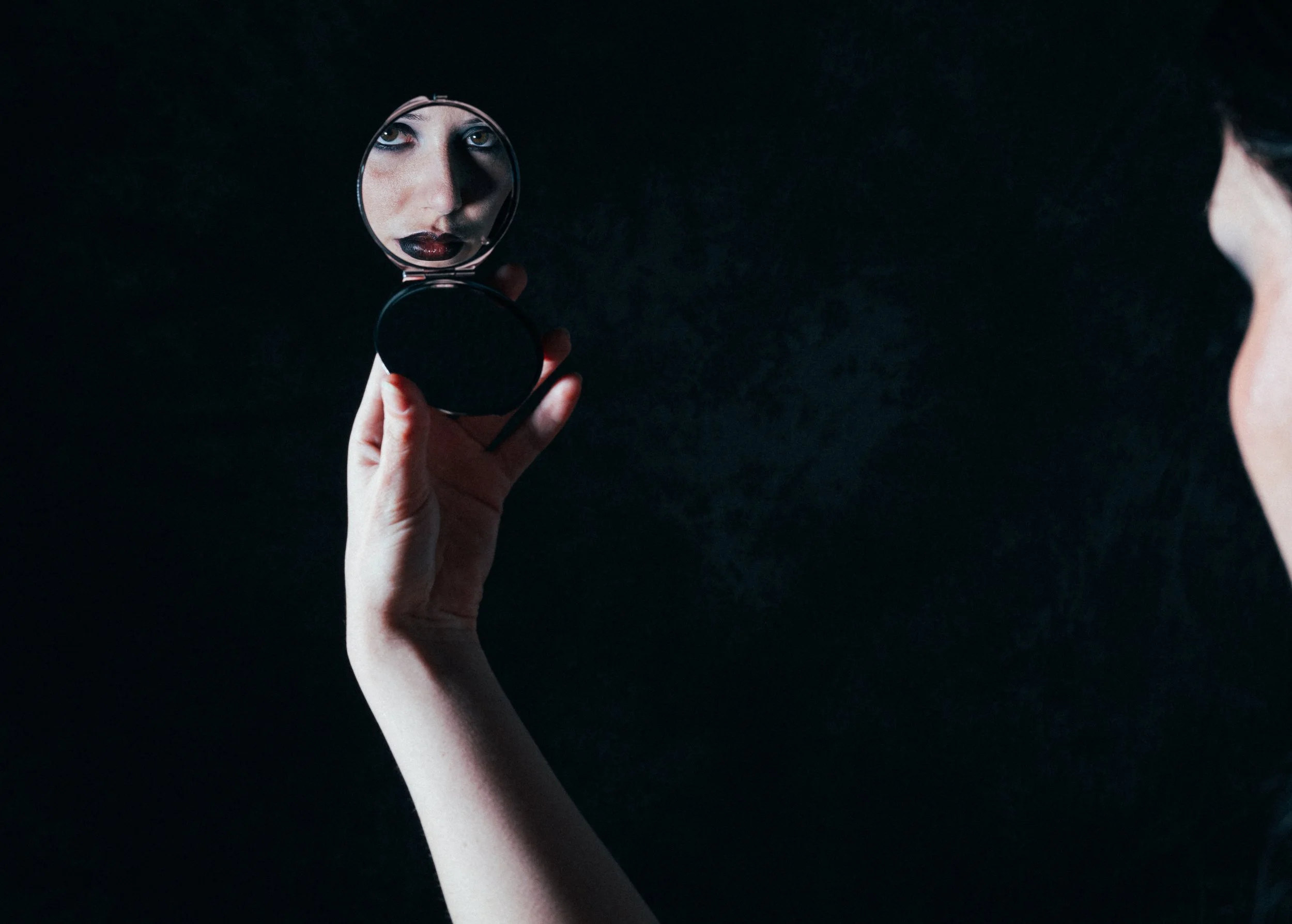 Une personne tient un miroir de maquillage vers le visage, reflet d'une femme avec un maquillage sombre.