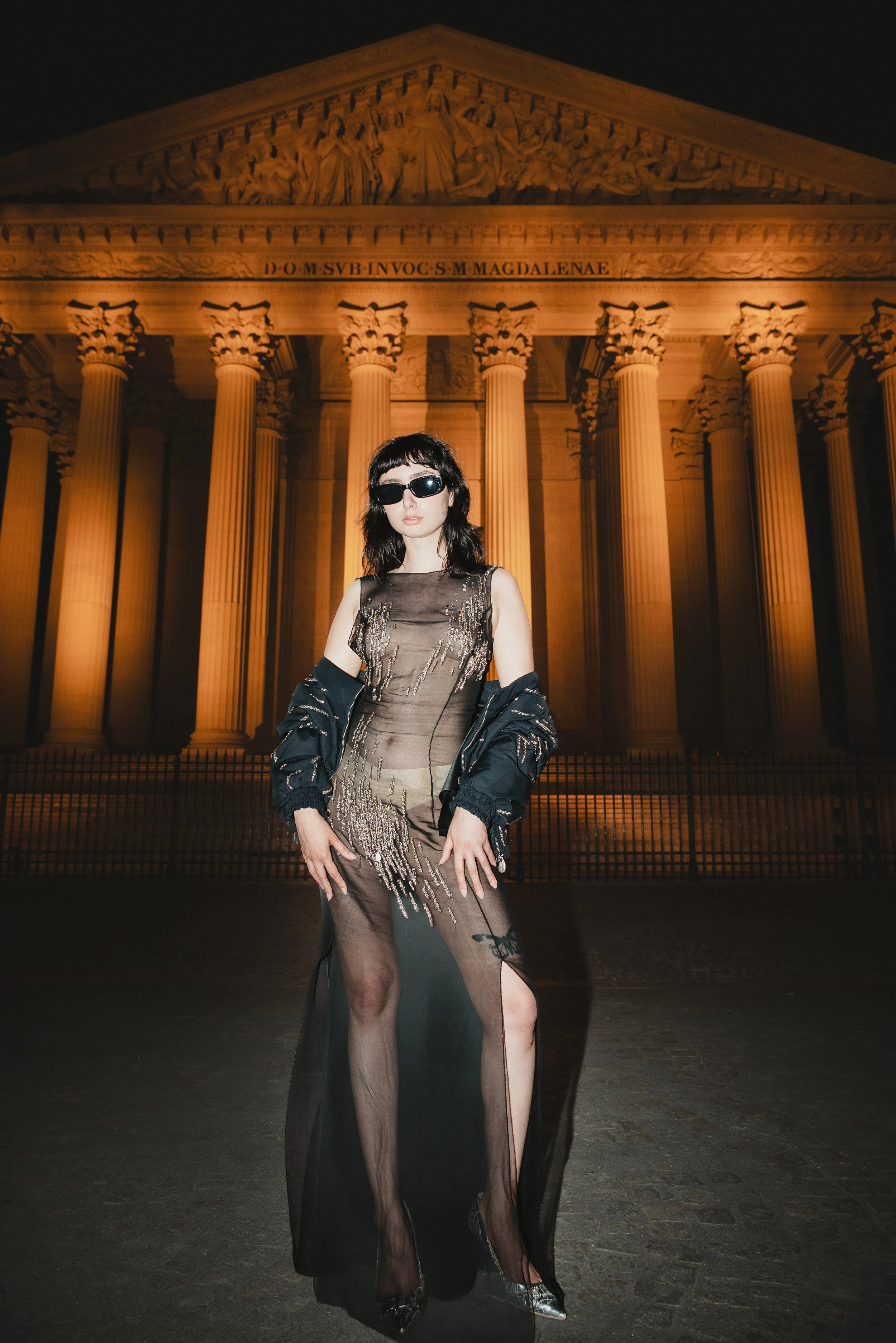 Une femme porte une robe transparente et des sandales à talons, avec une veste noire, devant un bâtiment historique éclairé la nuit.