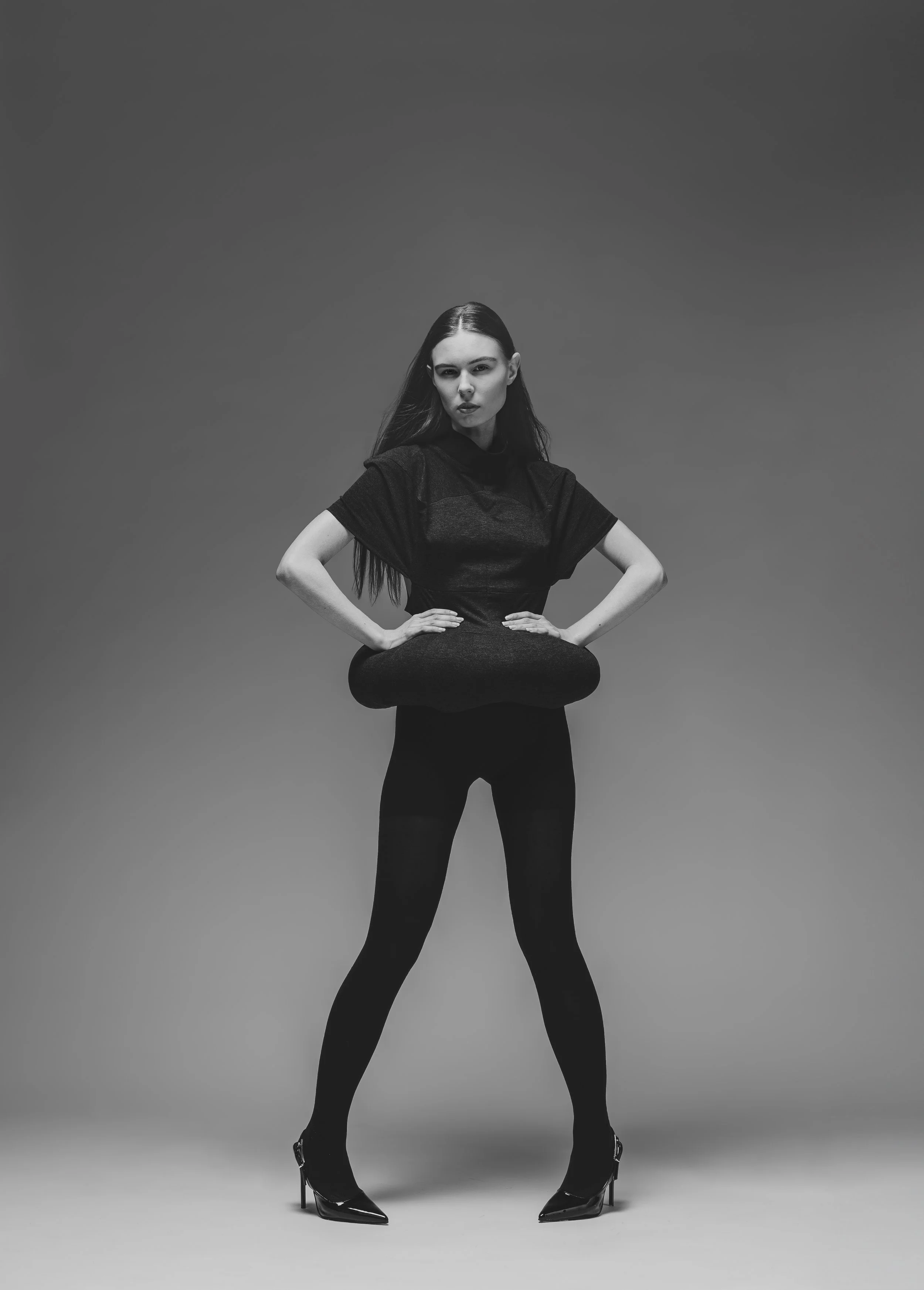 Une femme en robe noire et talons, avec un coussin, dans un studio avec fond gris.
