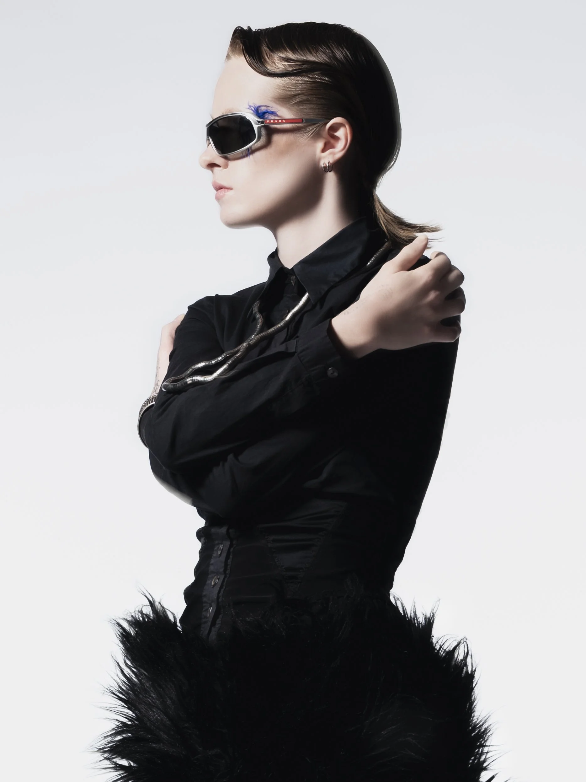 Une femme portant des lunettes de soleil de style moderne, une veste noire en tissu brillant, et un collier argenté, avec un fond blanc.