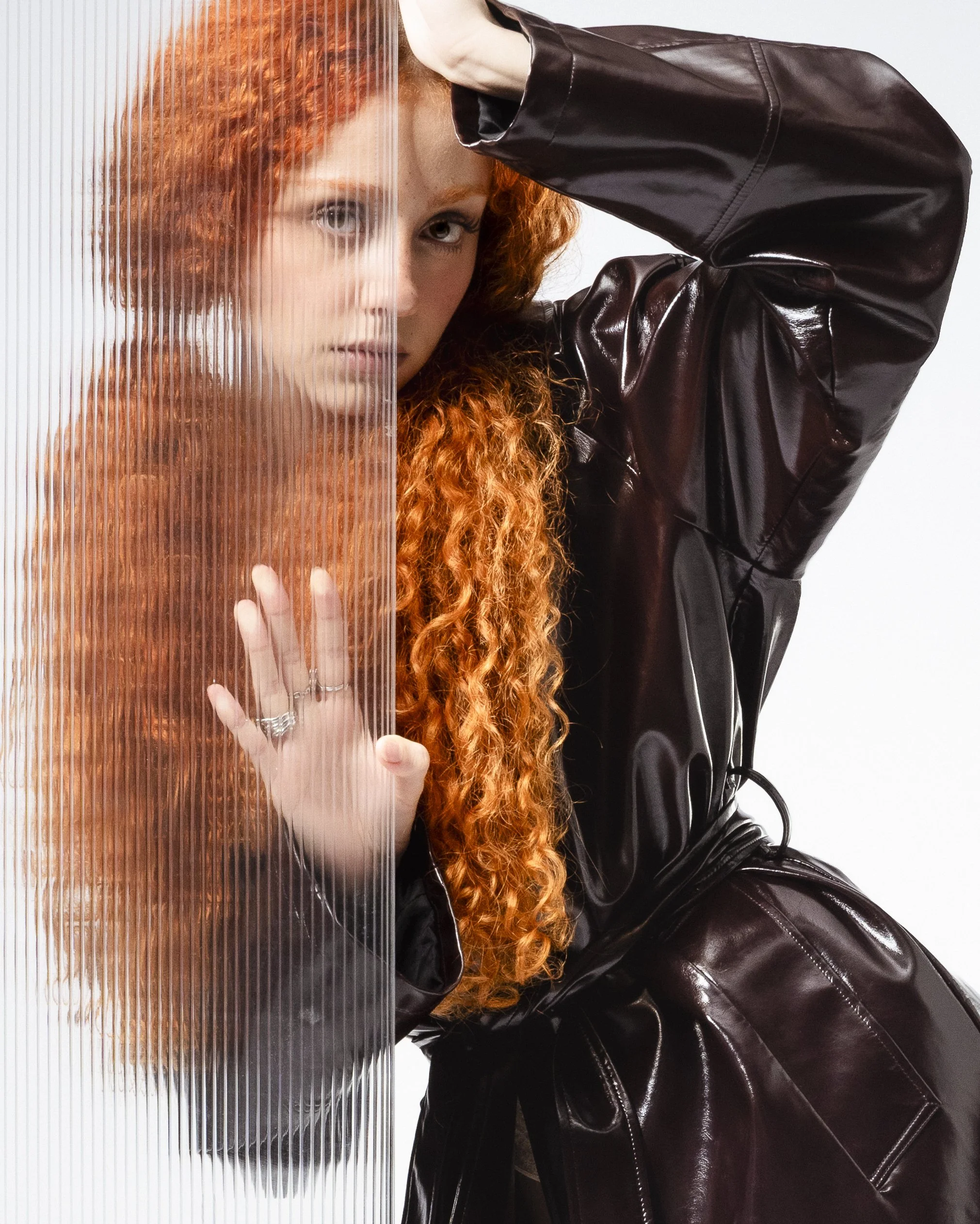 Femme aux cheveux roux, portant un manteau en vinyle noir, fendant un panneau translucide, regardant vers l'objectif.