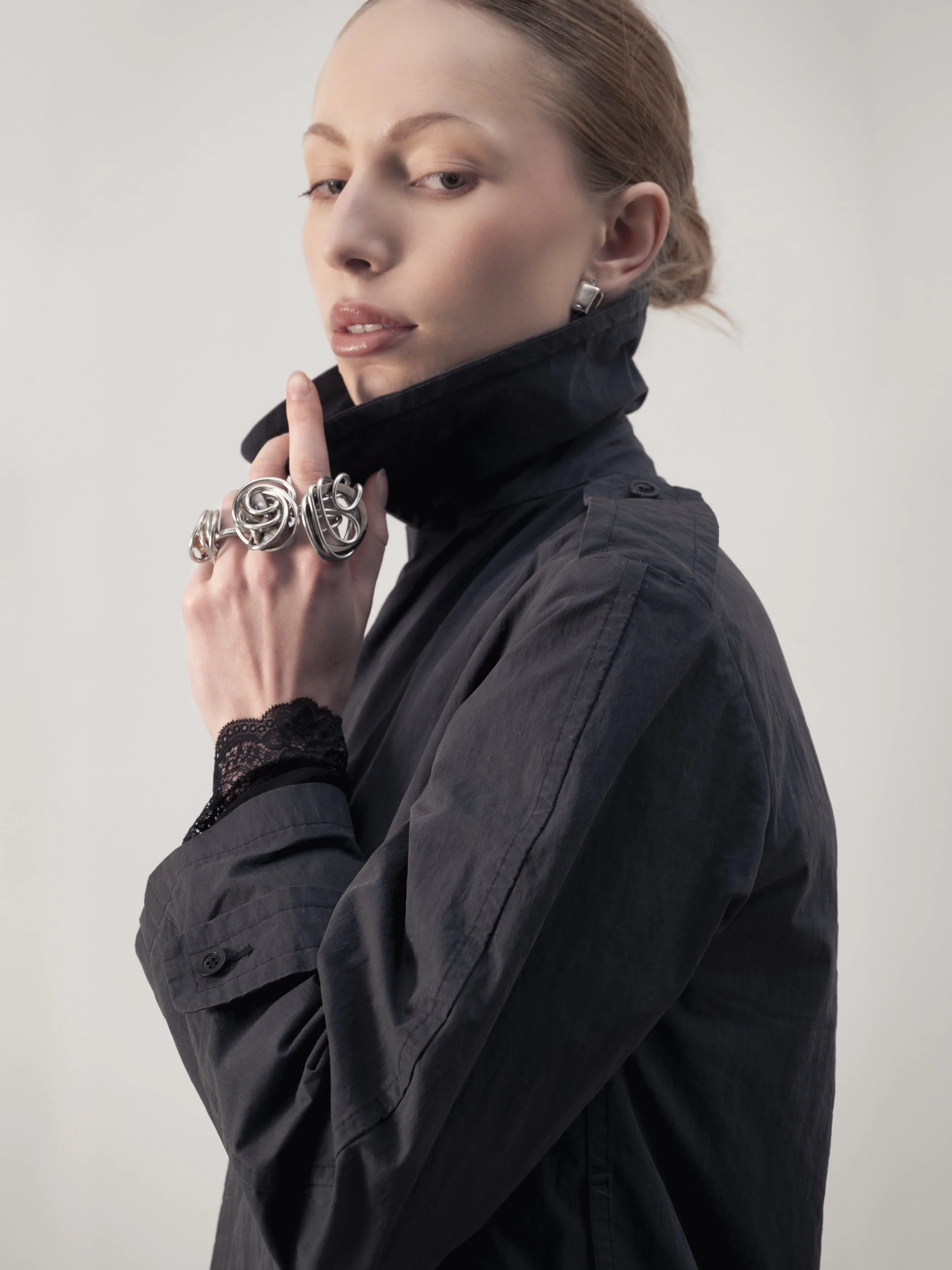 Jeune femme portant une veste noire, des bijoux en argent, et des bagues originales, posant avec une expression confiante contre un fond gris clair.