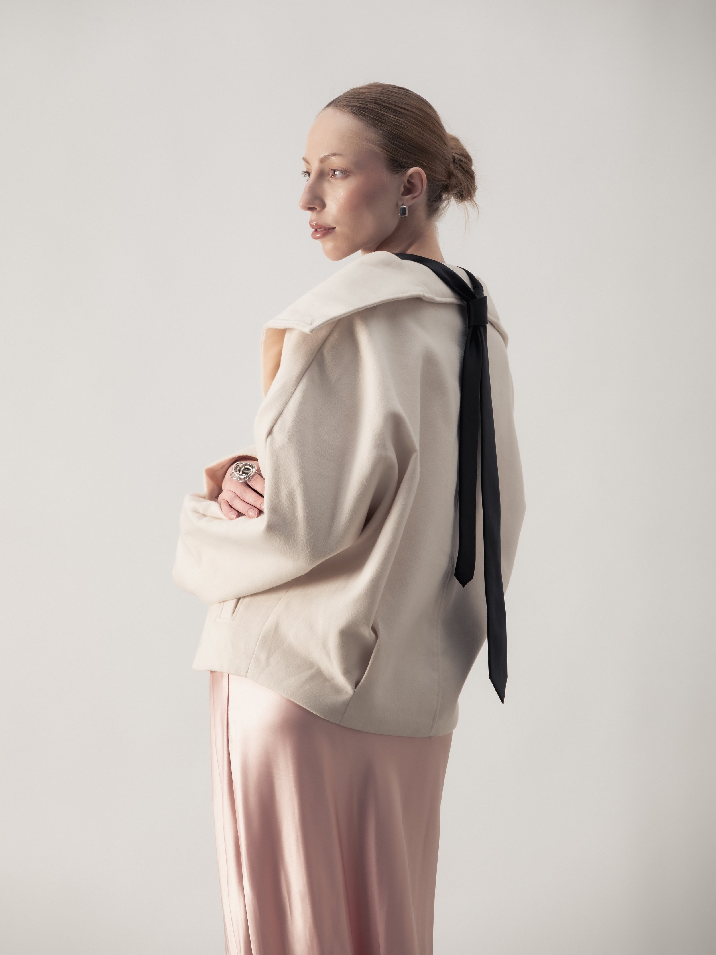 Une femme posant contre un fond blanc, portant un manteau beige, un pantalon rose en satin, et un foulard noir autour du cou.