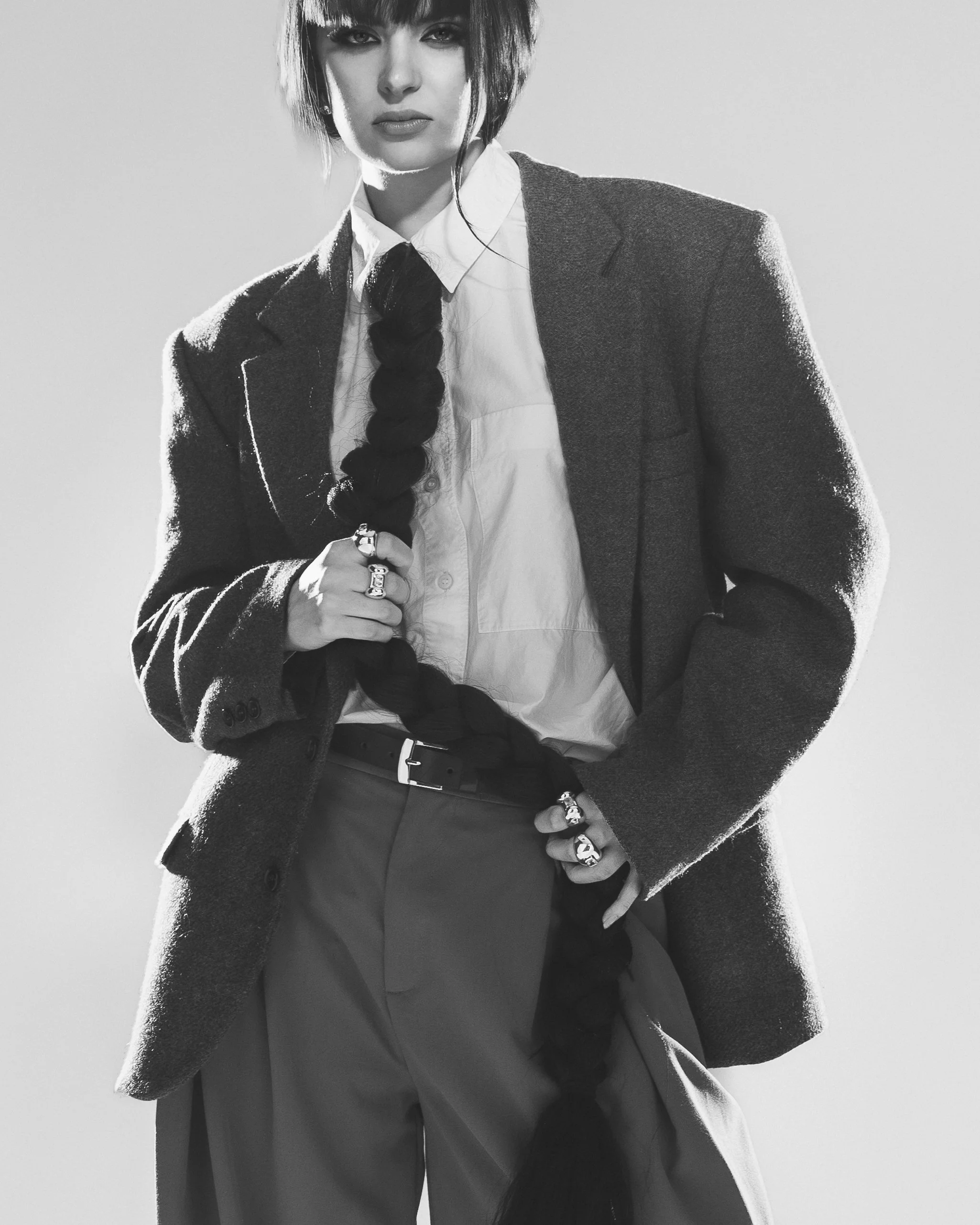 Une femme en costume avec une veste en tweed, une chemise blanche, un pantalon ample et des bijoux, posant pour une photo en noir et blanc.