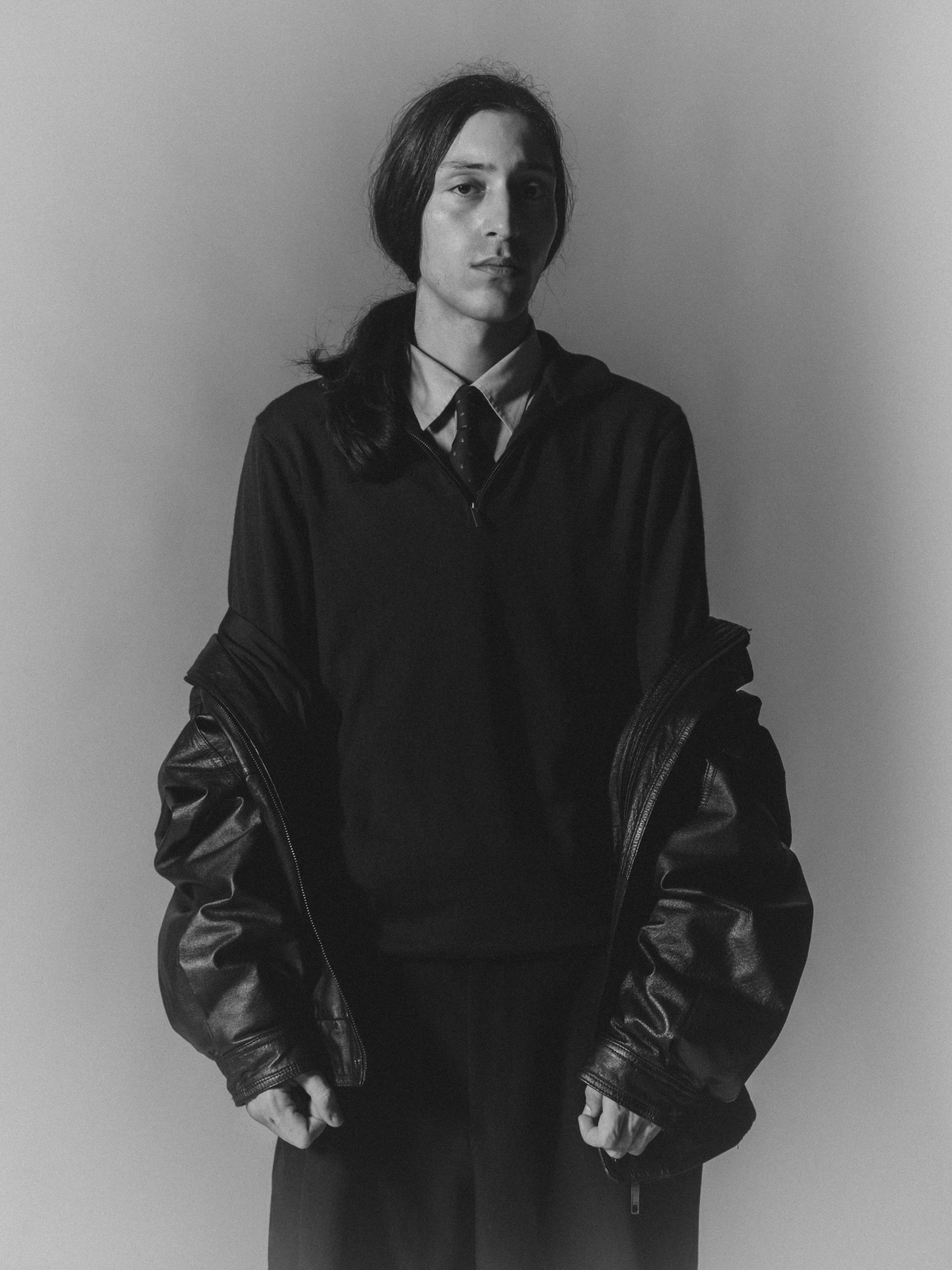 Une femme en noir et blanc, portant une veste en cuir sur une chemise, posant contre un mur clair.