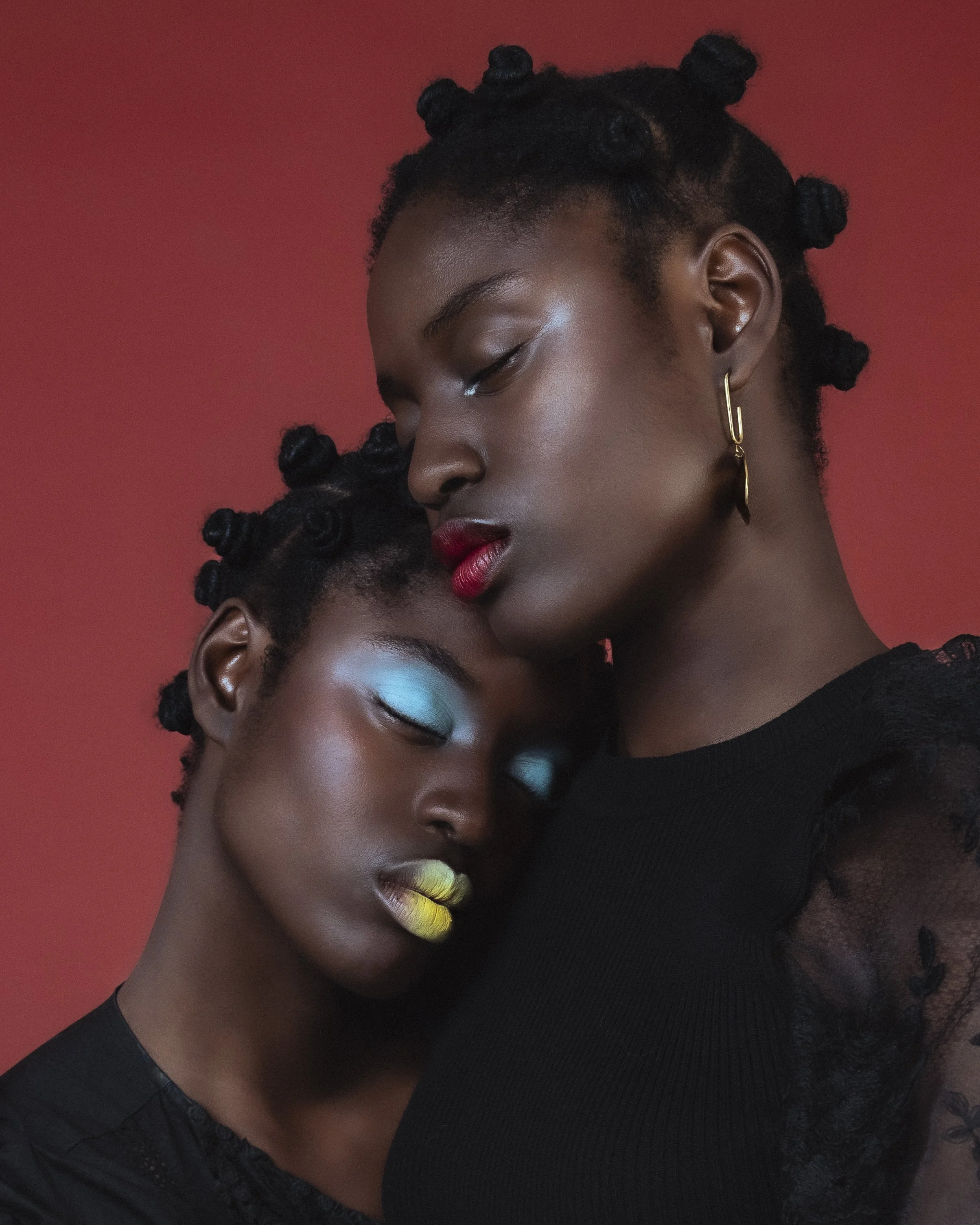 Deux femmes noires avec des cheveux tressés, maquillage coloré et vêtues de noir, se tiennent proche, avec un fond rouge.