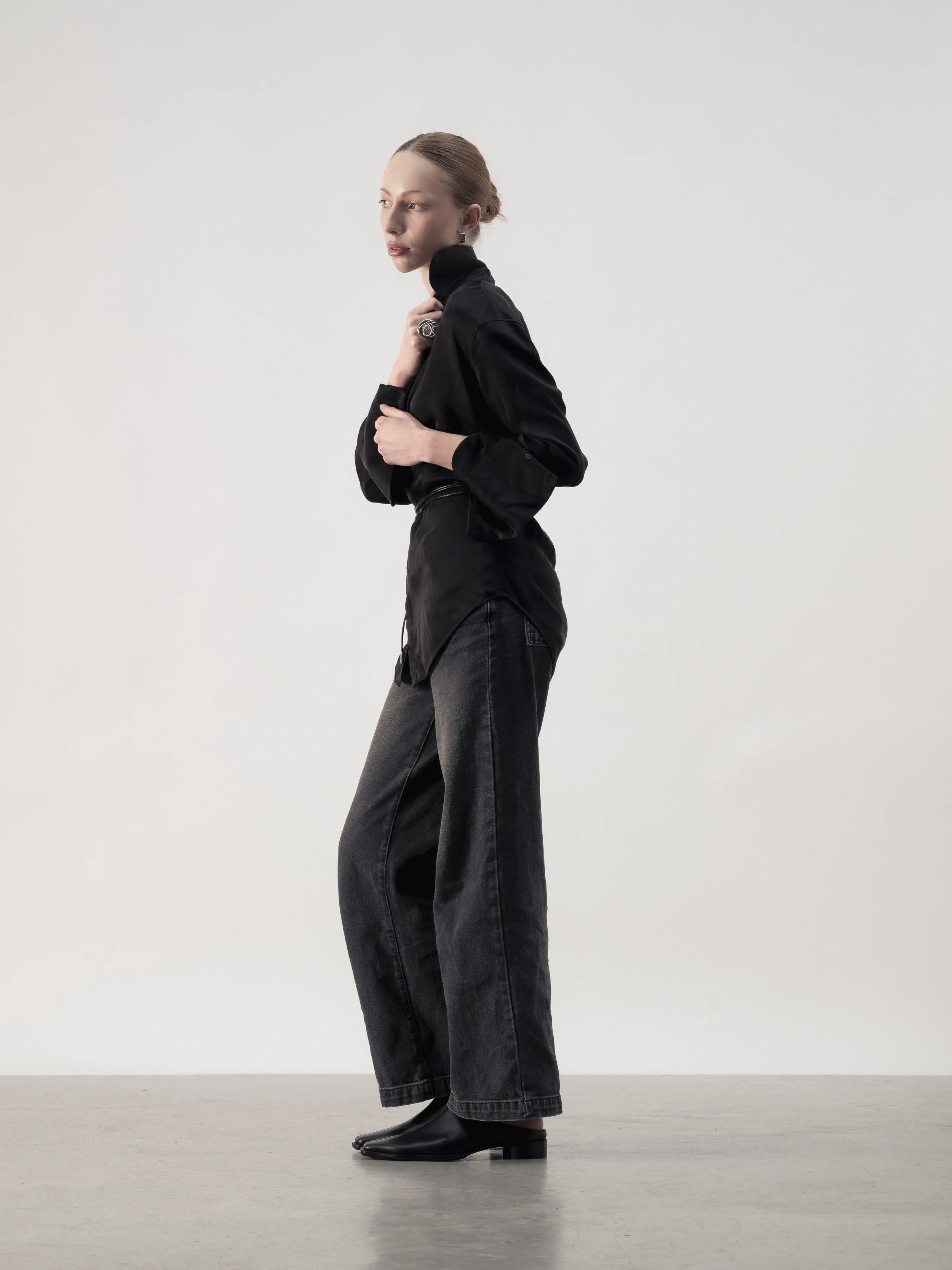 Femme avec pentalon noir ample, bottines noires et chemise noire à col montant, posant contre un fond blanc.