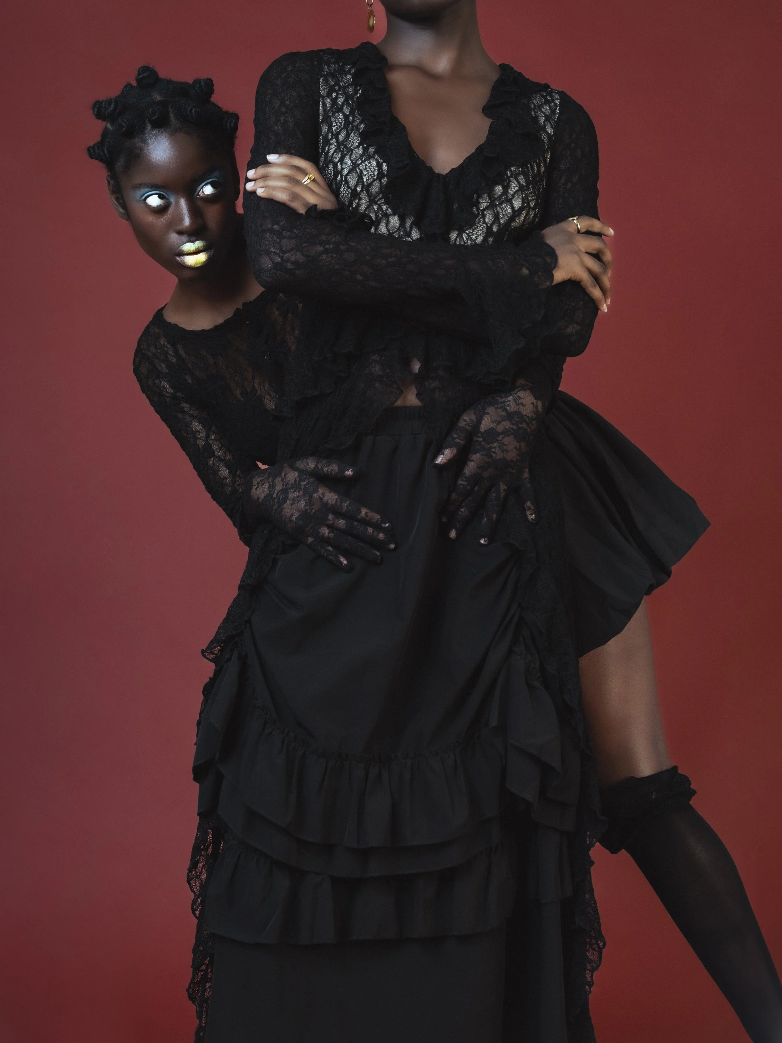 Deux femmes portant des tenues en dentelle noire posant contre un fond rouge. La femme en arrière-plan a la peau foncée, des boucles dans les cheveux et porte du maquillage audacieux. La femme en avant a aussi la peau foncée, avec des bras croisés et