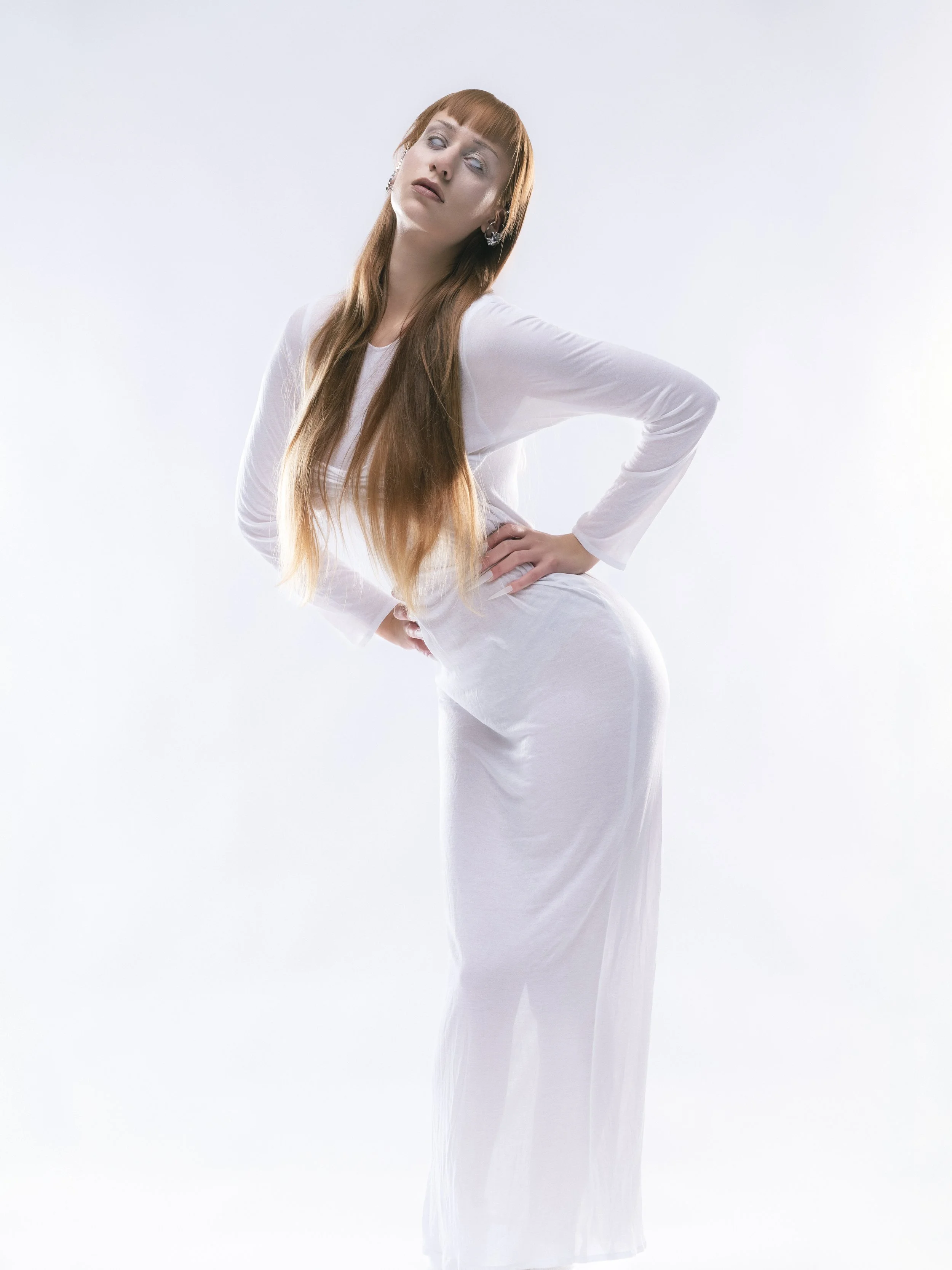 Une femme aux cheveux longs et roux, portant une robe blanche, pose devant un fond blanc, avec une expression de surprise ou de malaise.