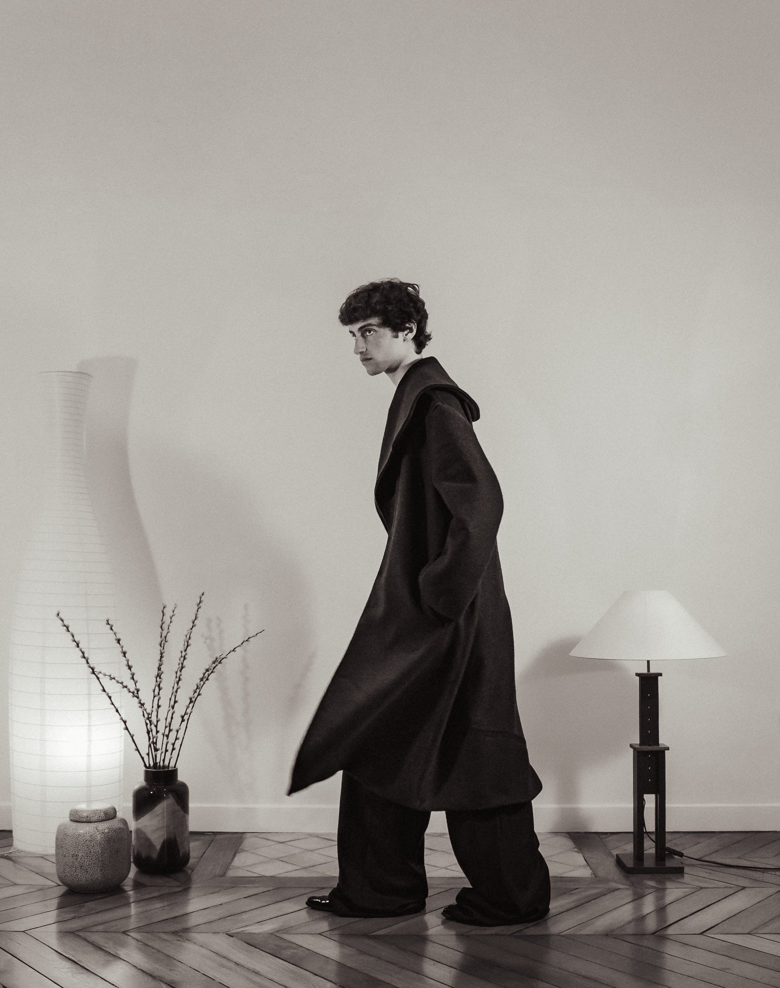 Un jeune homme en manteau noir, debout dans une pièce minimaliste, avec une lampe à côté et deux vases avec des branches, en noir et blanc.