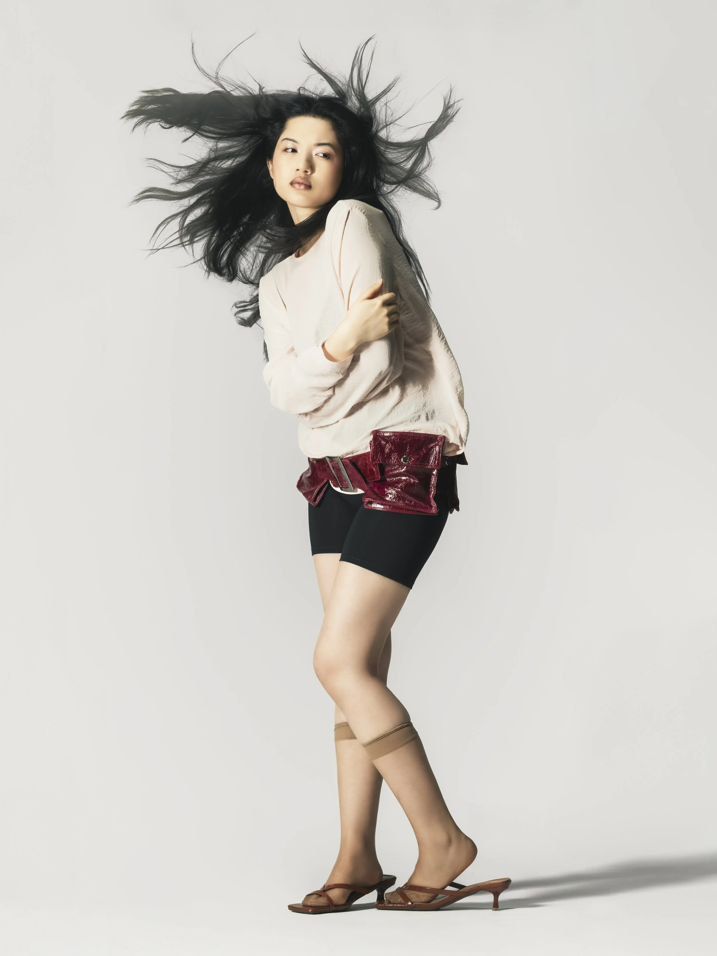 Une femme pose avec des cheveux noirs flottants, portant un haut clair, un pantalon court, des bas beiges, des sandales à talons et une ceinture rouge en plastique brillant.