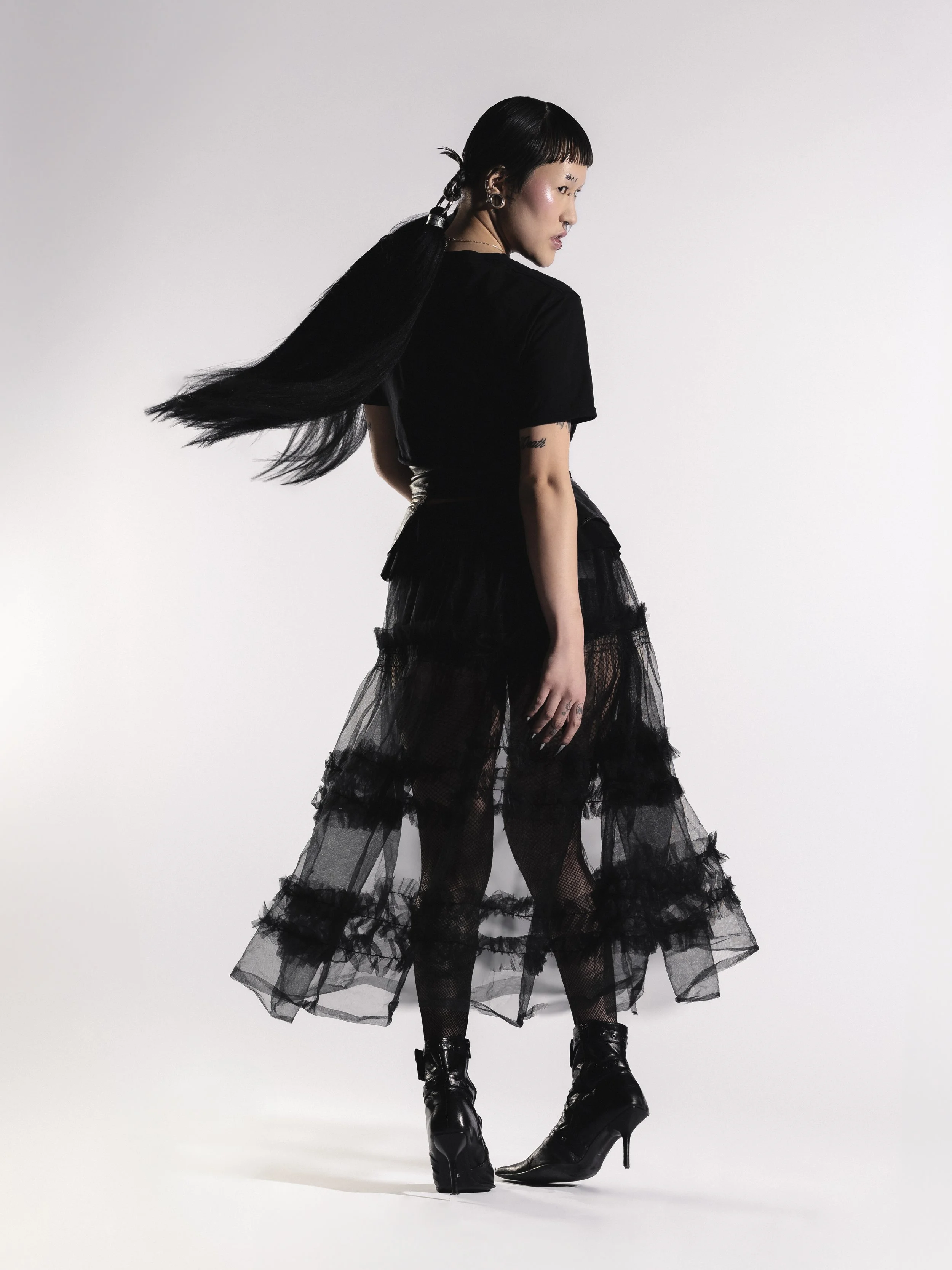 Une femme portant un ensemble noir avec une jupe en tulle et des bottes à talons, posant contre un fond blanc.