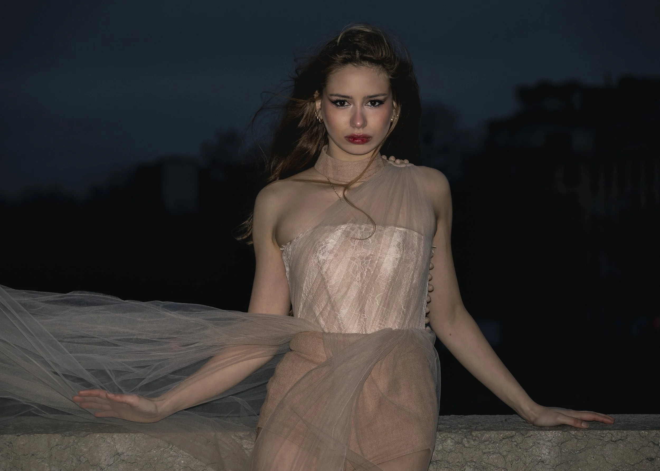 Jeune femme posant près d'un mur la nuit, portant une robe en tulle beige, avec un maquillage marqué et des boucles d'oreilles.