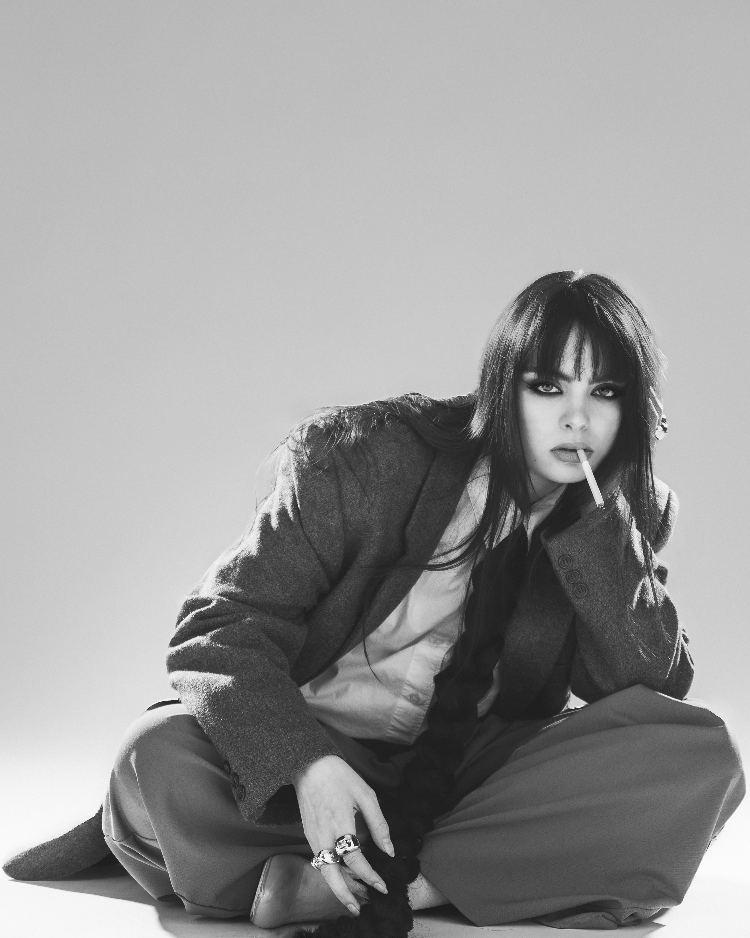 Une femme avec des cheveux longs et noirs, portant une veste en tissu et un pantalon ample, tenant un cigarette dans la bouche, assise par terre en position décontractée, dans une photographie en noir et blanc.