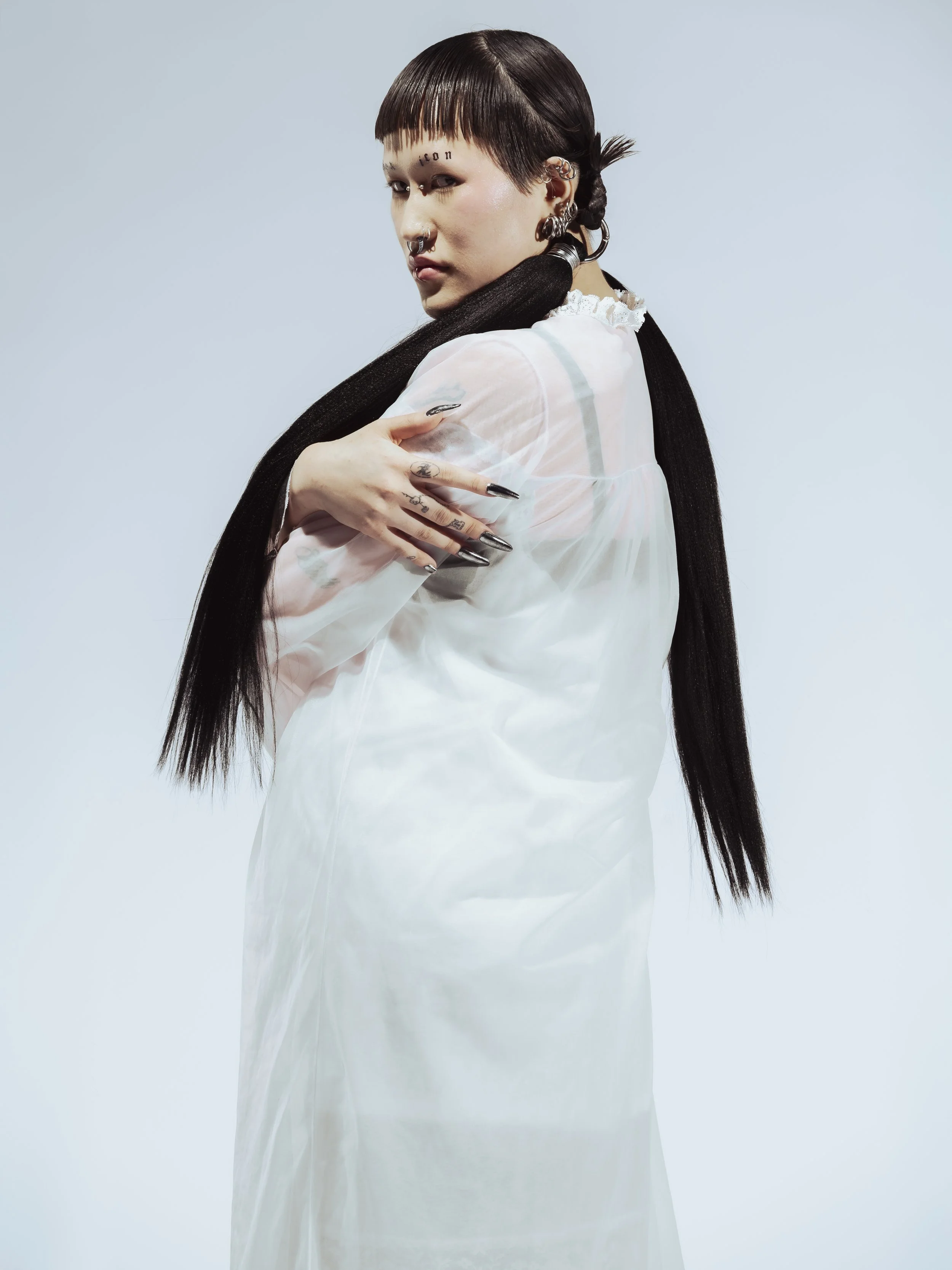 Une femme avec des tatouages visibles, portant une robe blanche translucide, avec de longs cheveux foncés et coiffés en tresses, pose contre un fond gris clair.