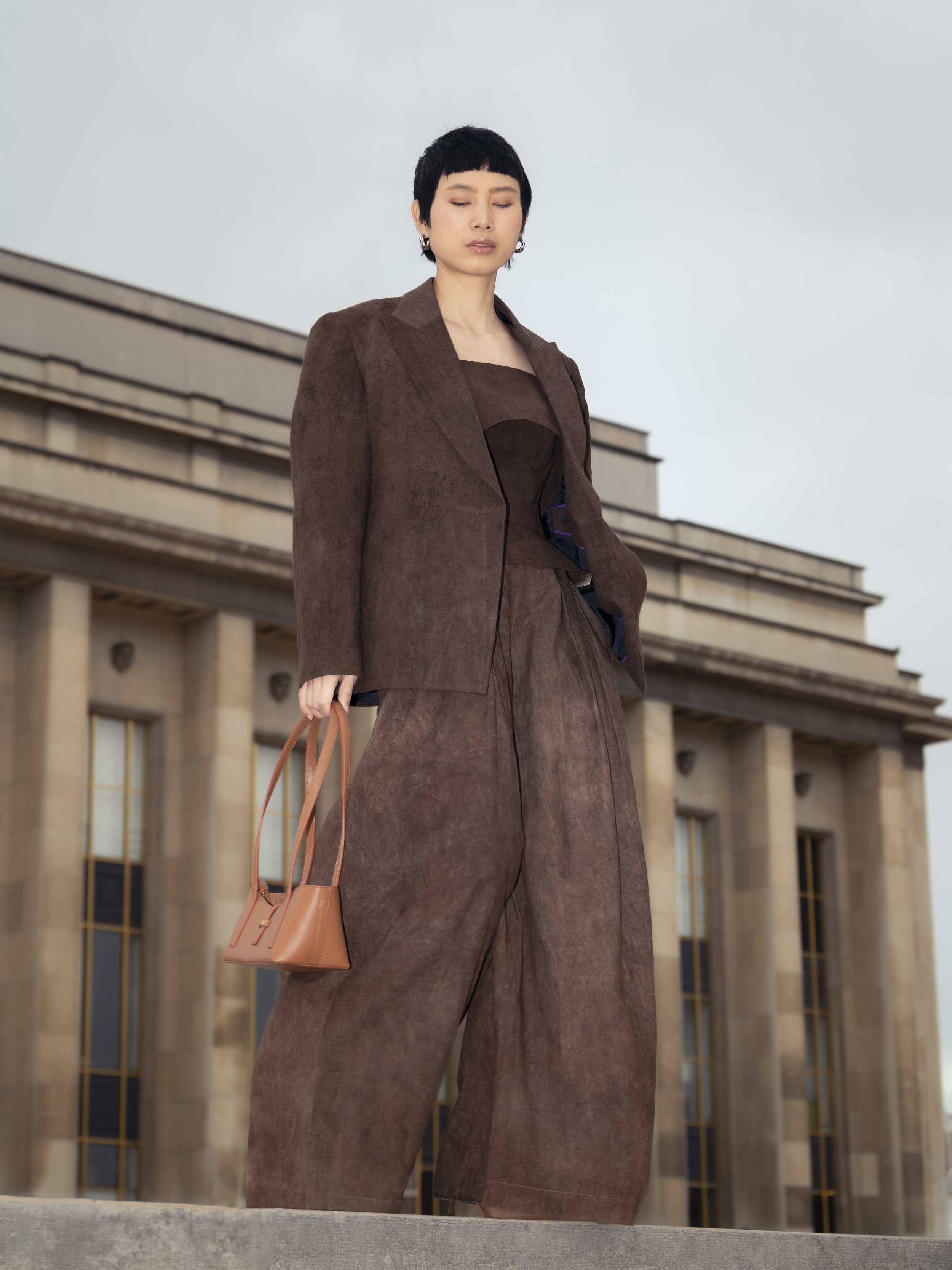 Femme avec une tenue en cachemire marron, portant un sac à main beige, devant un bâtiment classique en pierre