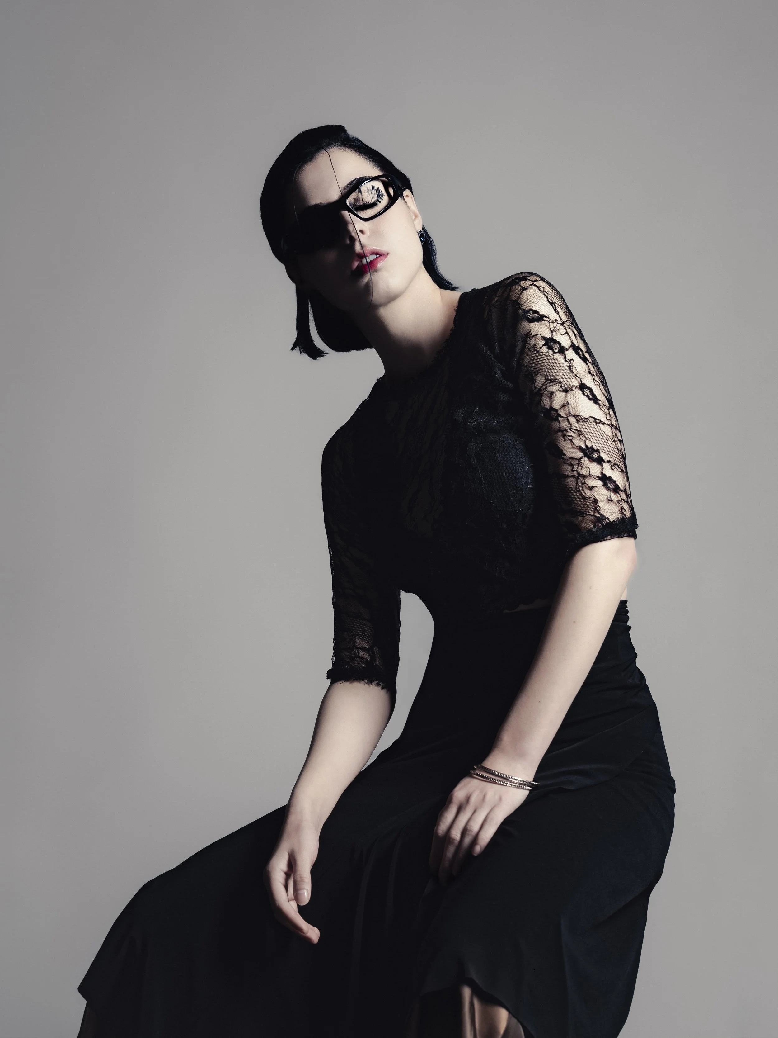 Une femme élégante portant une robe noire en dentelle, lunettes noires, avec des cheveux courts noirs, assise sur une surface, contre un fond neutre.