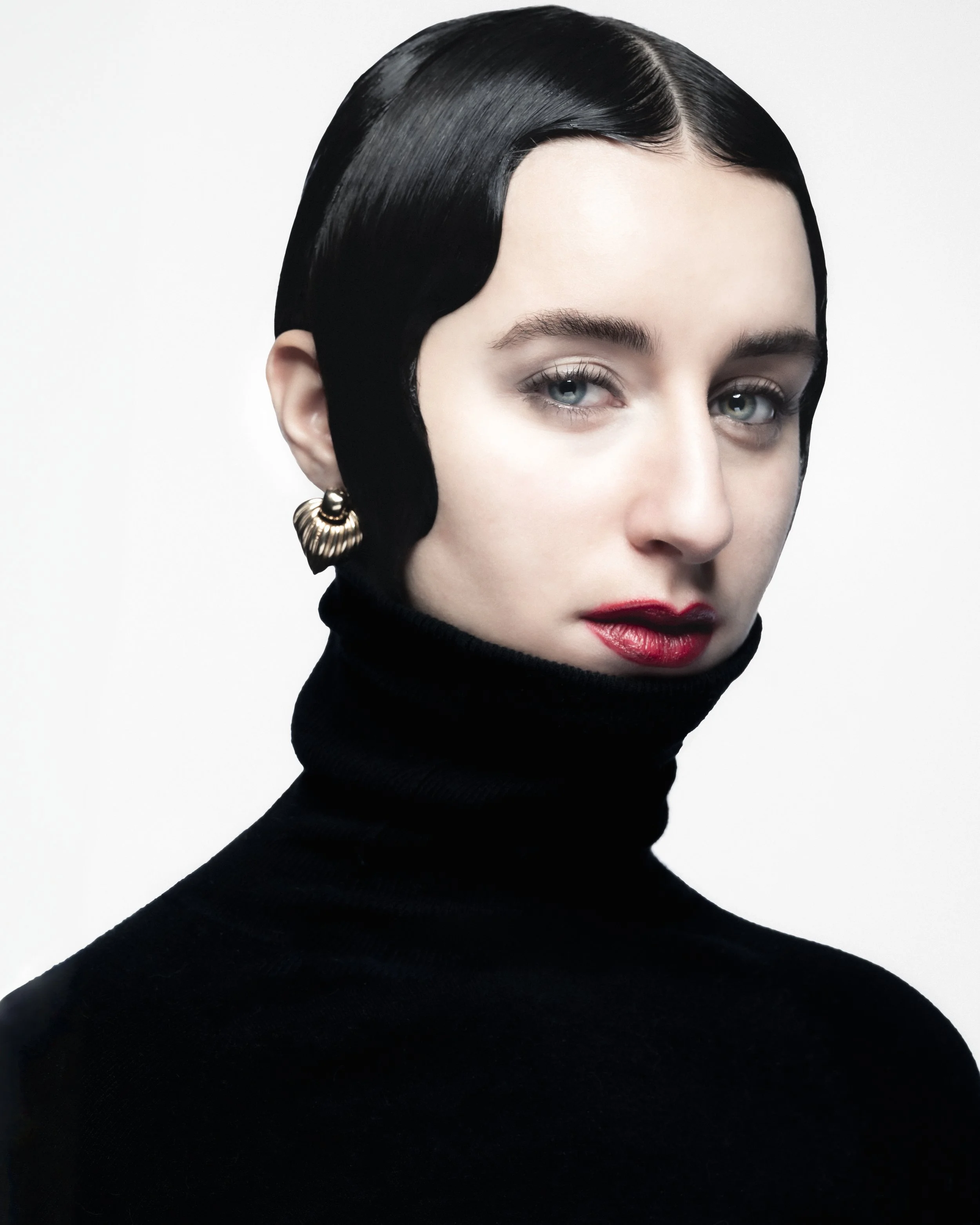 Femme avec cheveux courts, noir et brillant, portant un col roulé noir et une boucle d'oreille en forme de coquillage doré.