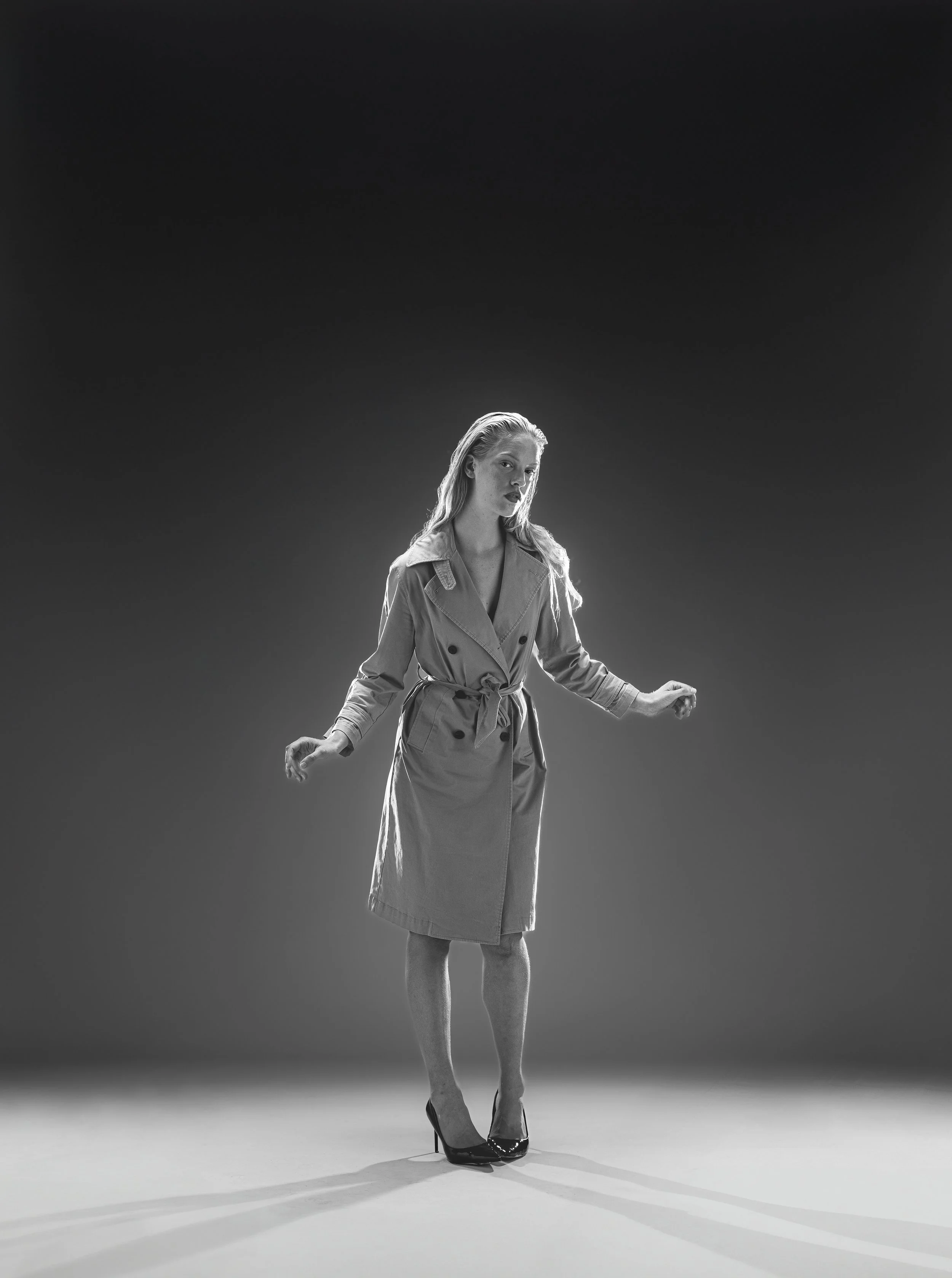 Une femme en trench coat et talons hauts se tient sur un fond sombre, éclairée sous un éclairage directionnel en noir et blanc.