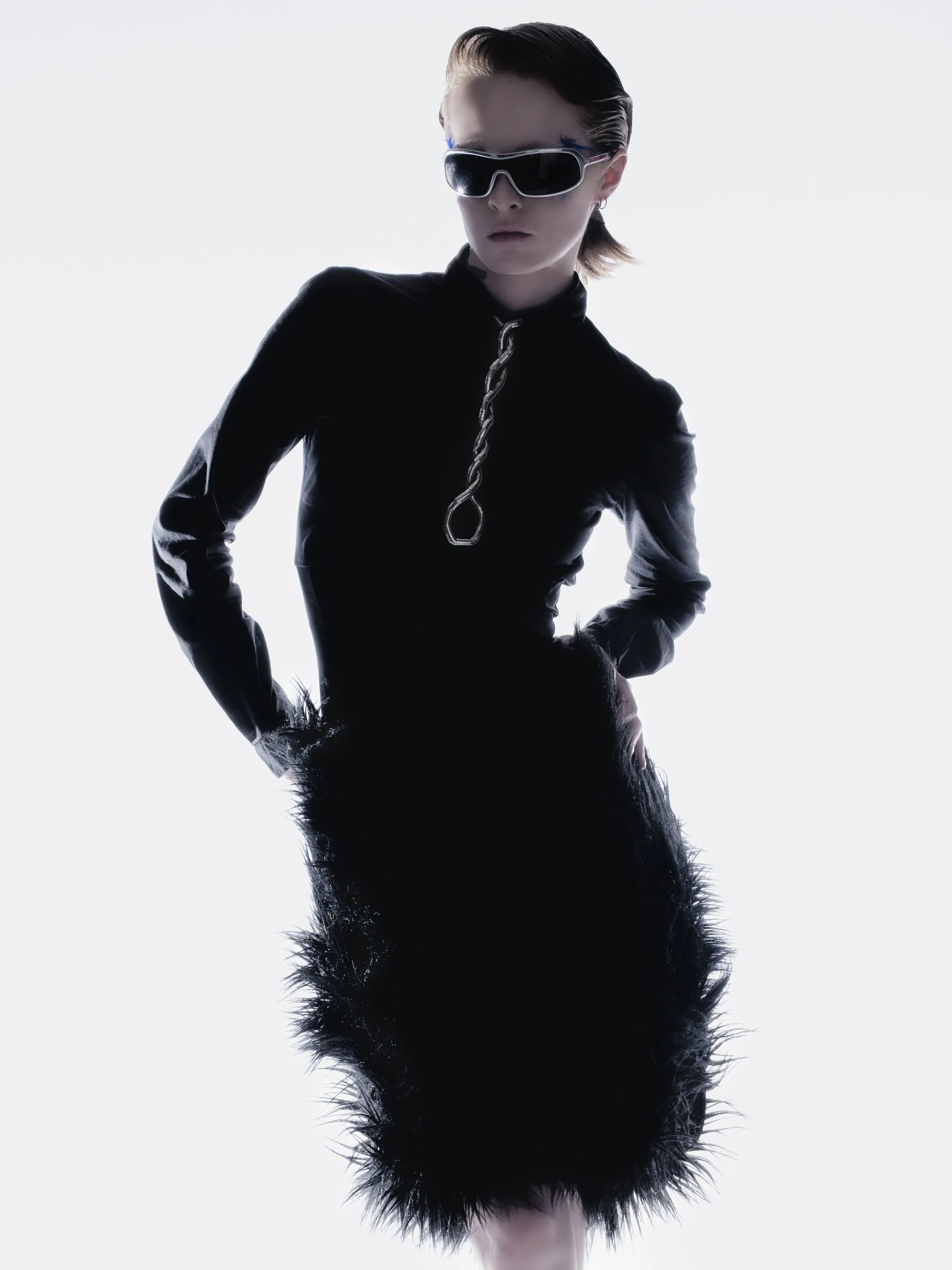 Une femme portant des lunettes de soleil et une robe noire avec un col en chaîne, pose avec les mains sur les hanches devant un fond clair.