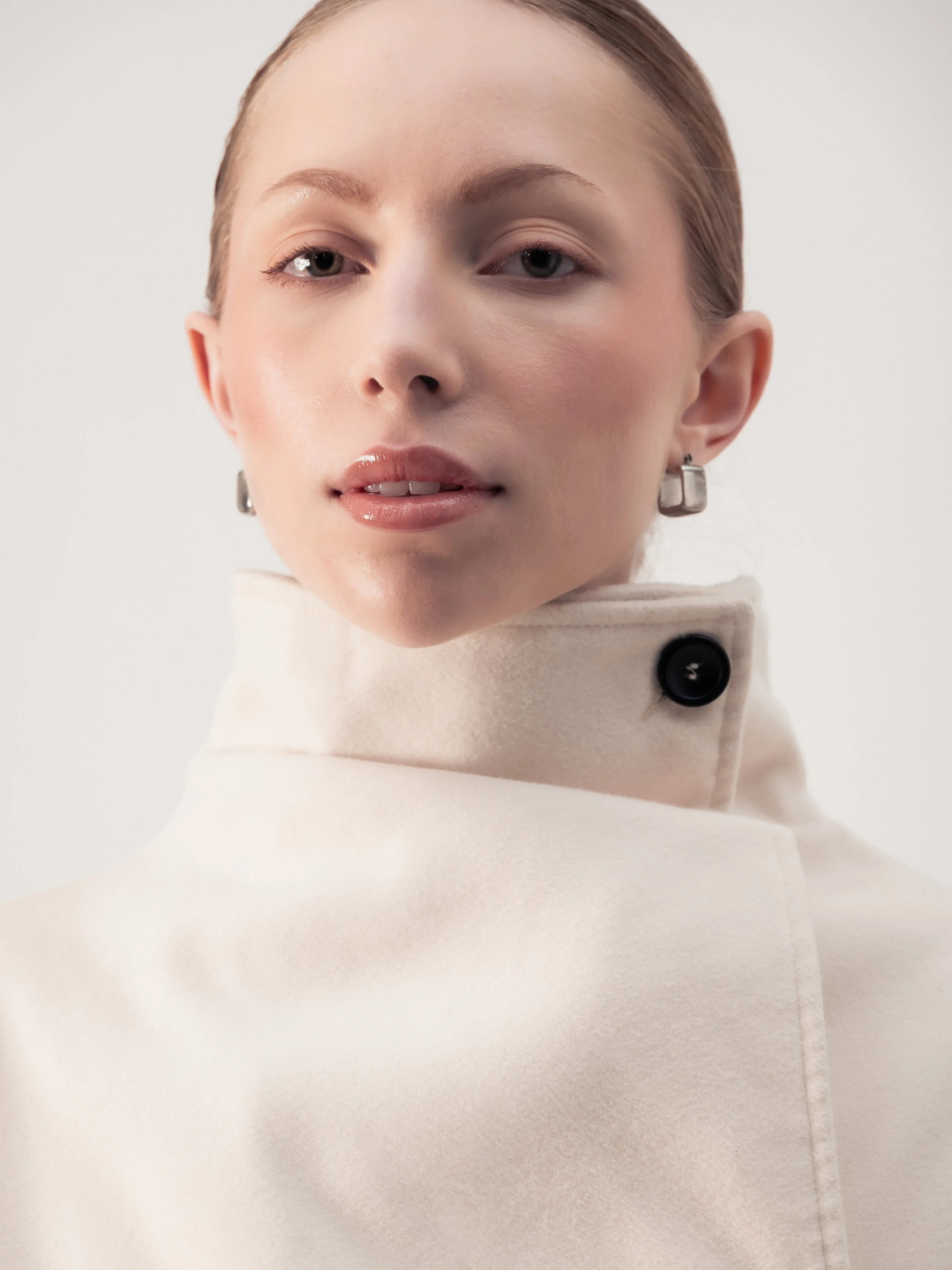 Portrait d'une femme avec un maquillage naturel, portant un manteau blanc avec un col haut et un bouton noir, portant des boucles d'oreilles carrées en argent.