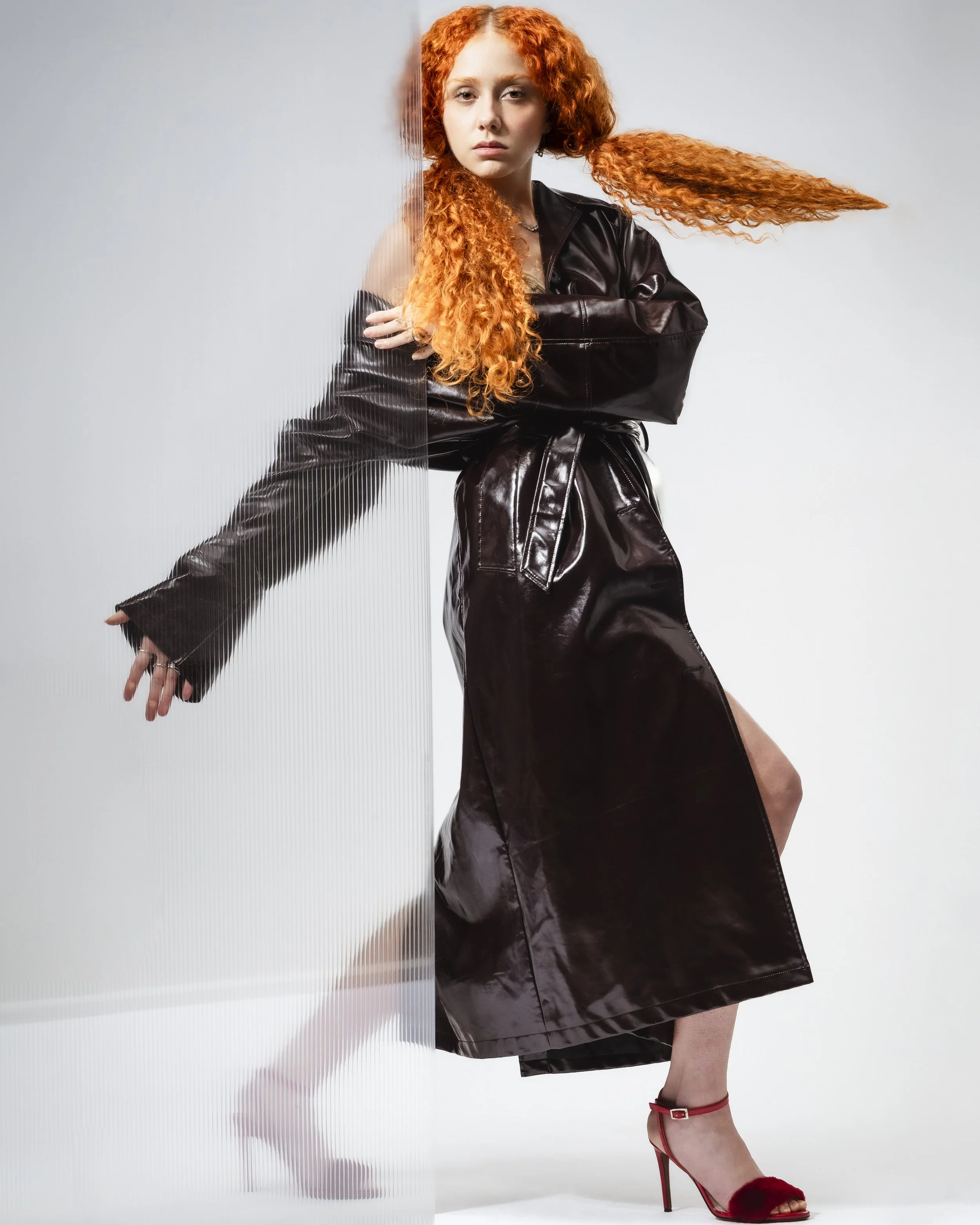 Femme aux cheveux roux portant un manteau en cuir noir, avec une jambe visible en haut de la robe. Elle porte des chaussures à talons rouges avec des détails en fourrure rouge.