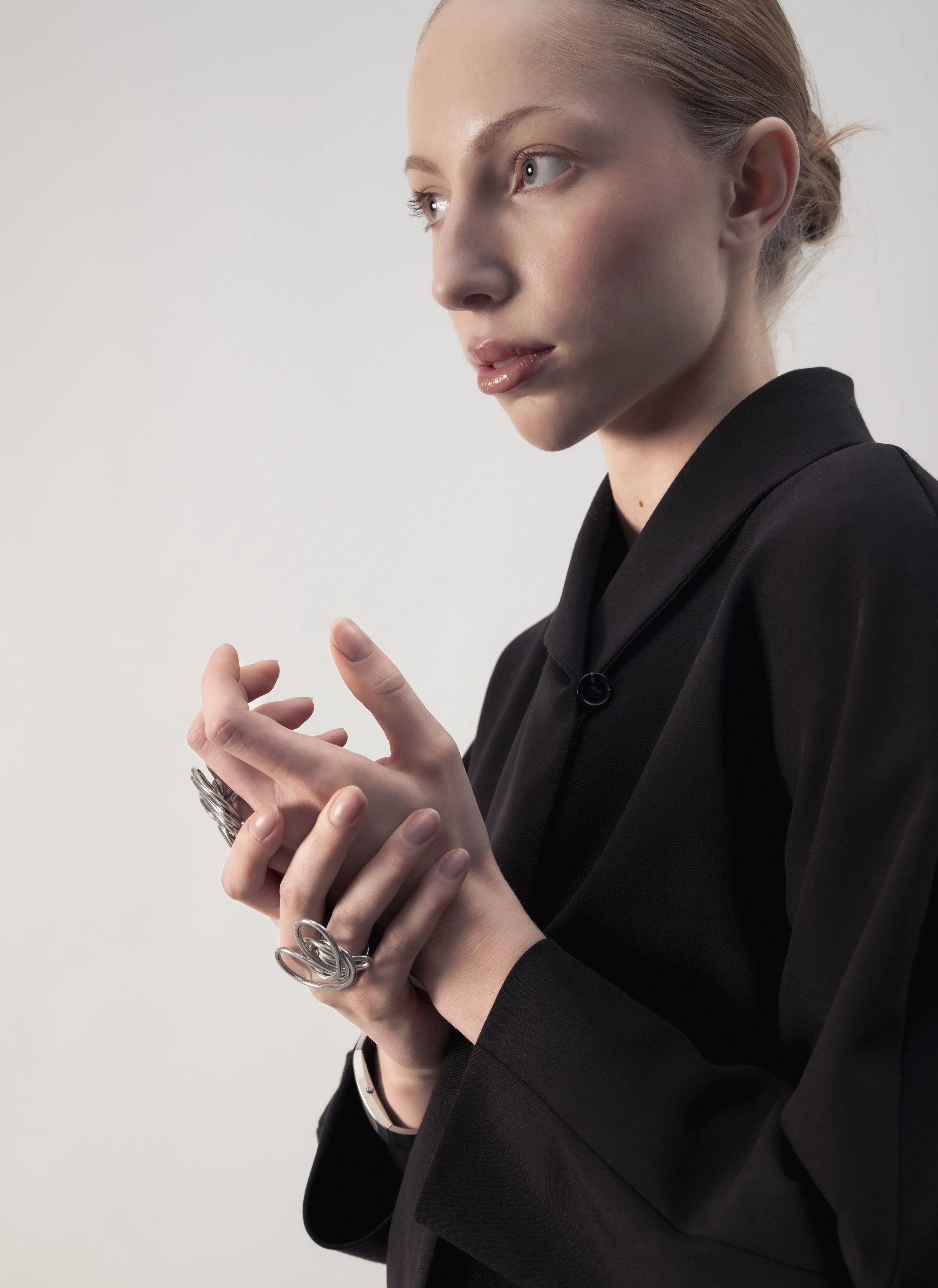 Une jeune femme portant un costume noir, avec des anneaux en métal à ses doigts, regarde vers la gauche, dans un contexte neutre et bien éclairé.