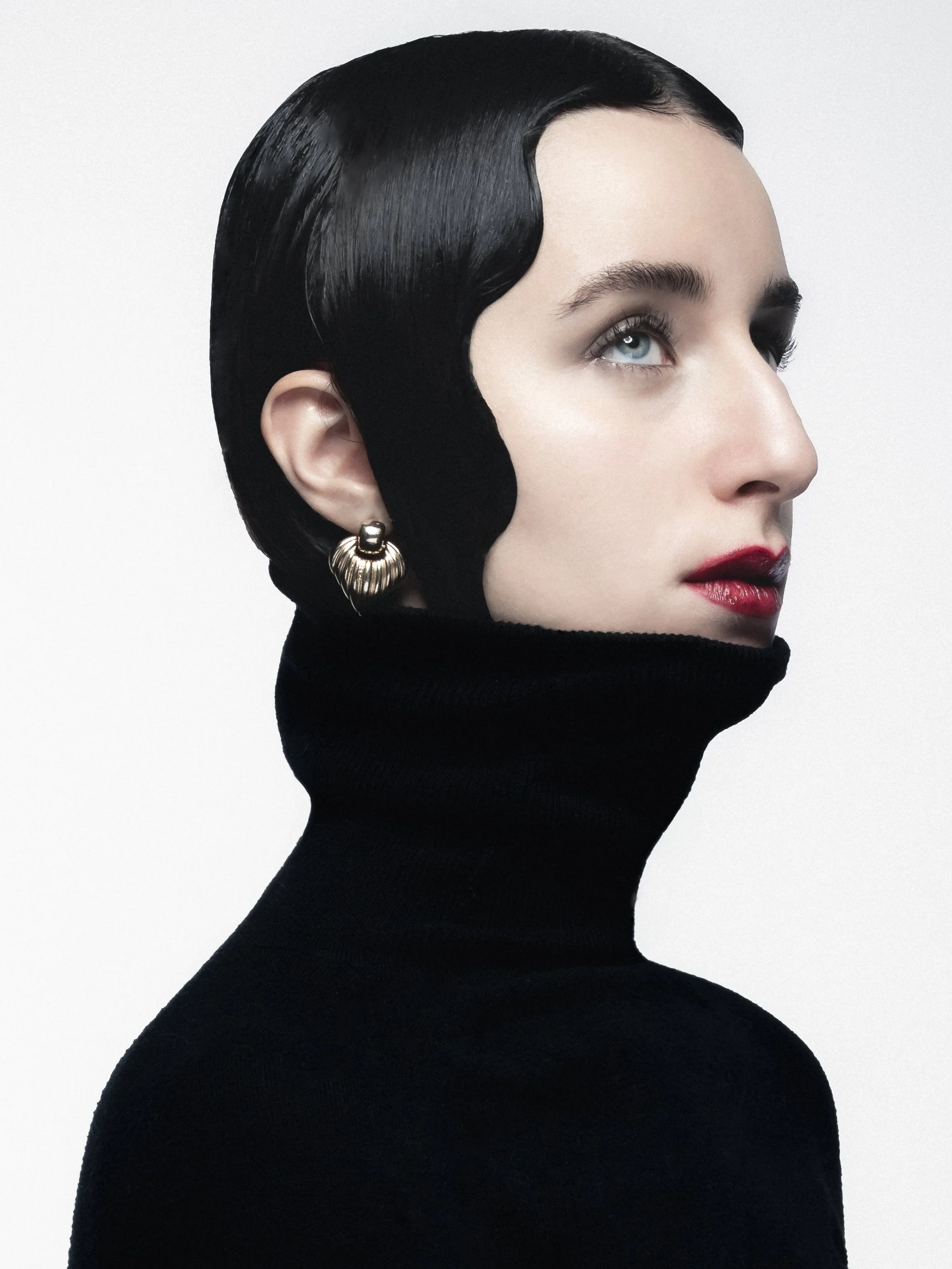 Une femme au visage clair, avec des cheveux noirs lisses, porte un haut noir à col roulé, des boucles d'oreilles en or et a des lèvres rouges, regardant légèrement vers la droite.