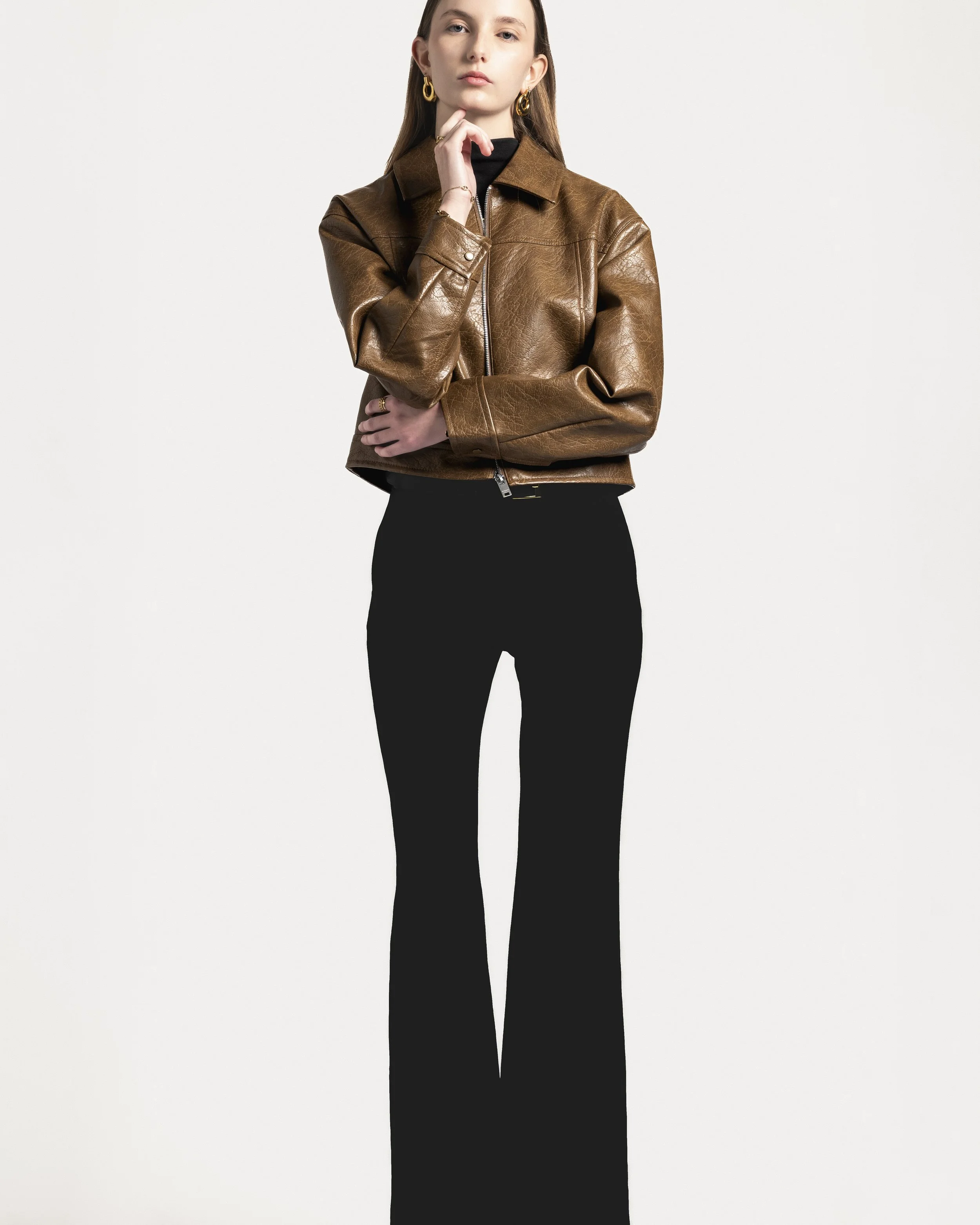 Femme portant une veste en cuir marron et un pantalon noir, posant contre un fond blanc.