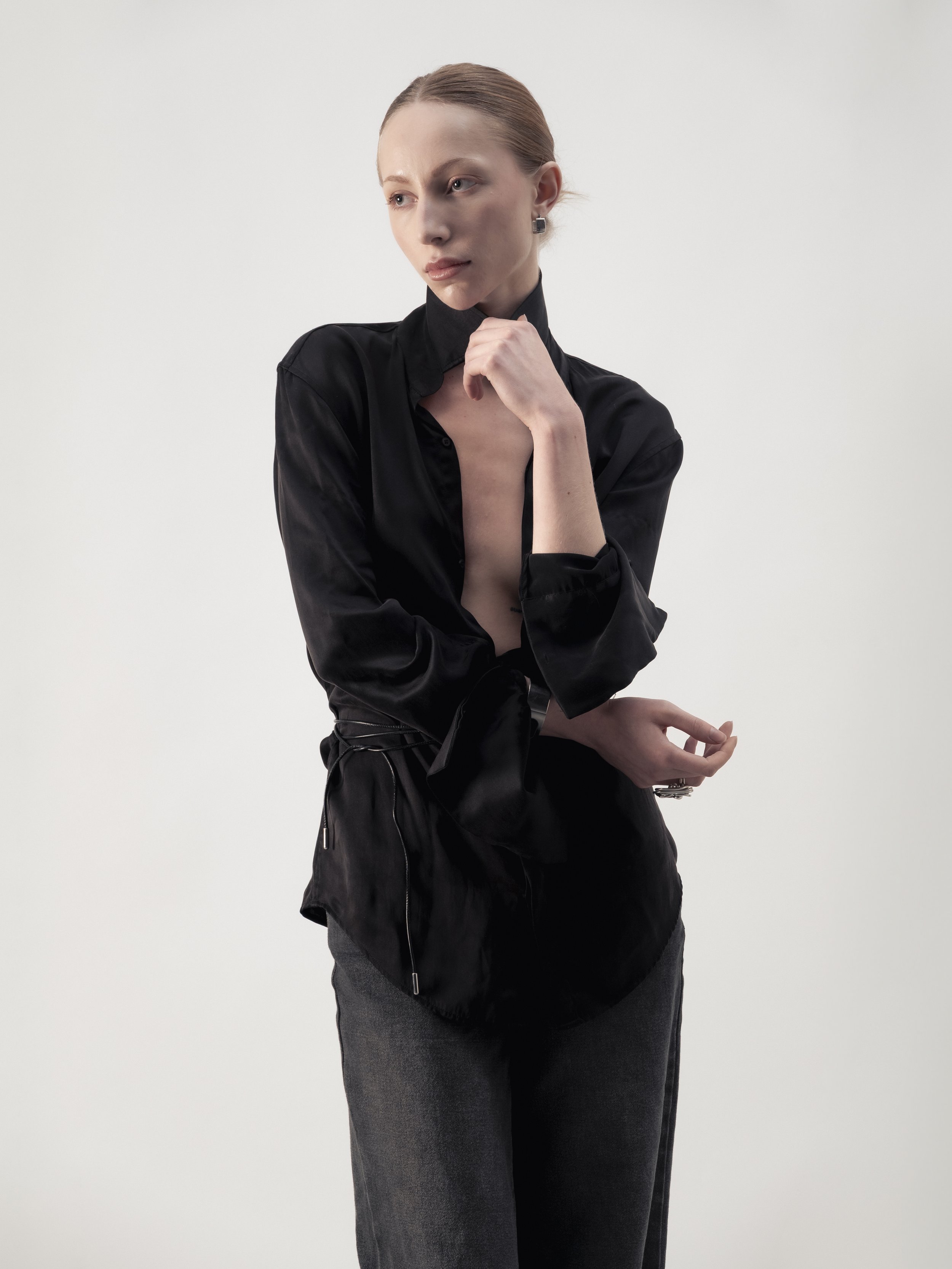 Une jeune femme avec des cheveux roux attachés, portant une blouse noire et un pantalon gris foncé, pose contre un fond blanc.