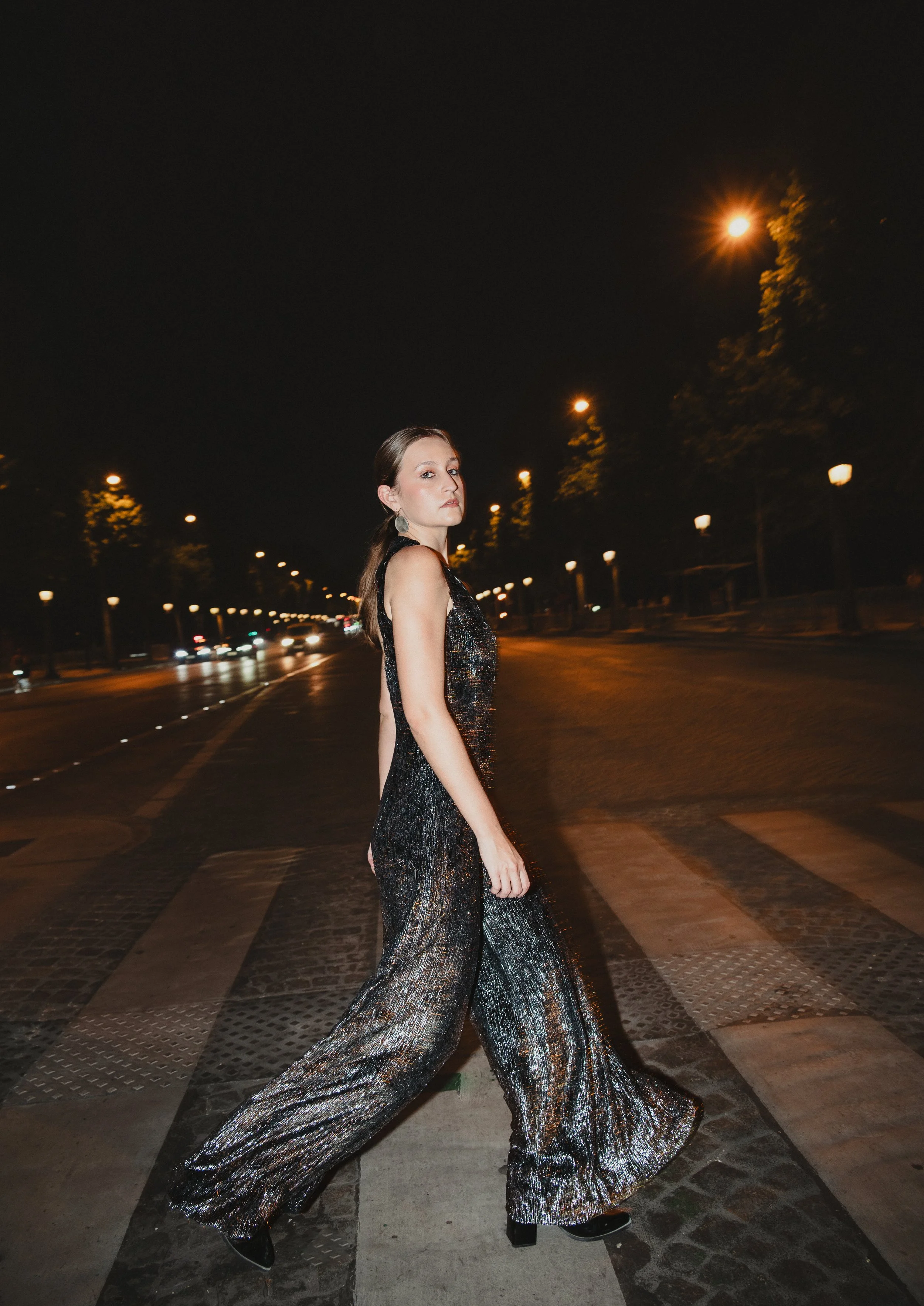 Femme portant une robe longue en soie noire à sequins, traversant un passage piéton la nuit, avec des lampadaires illumine la rue.