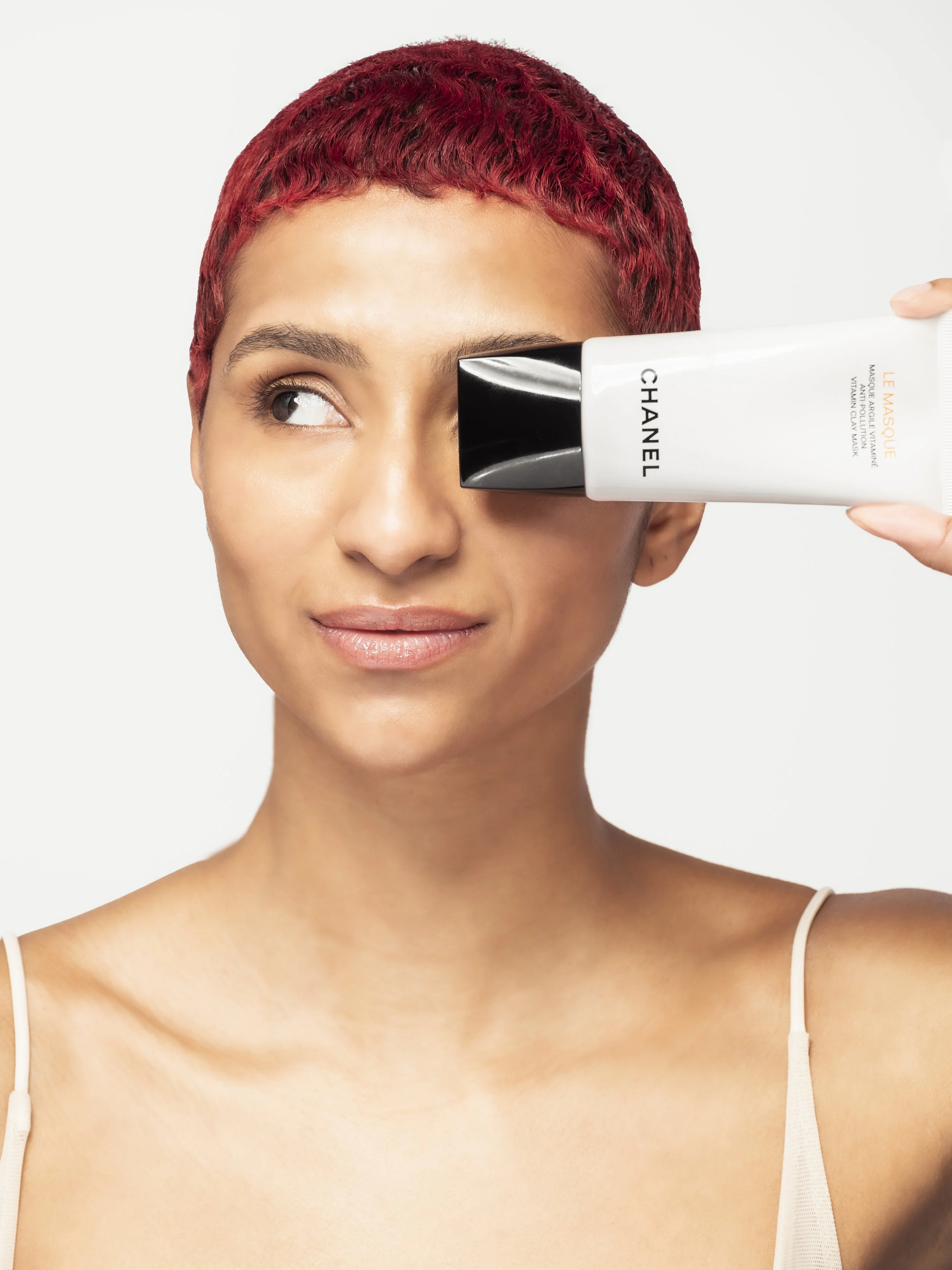 Femme avec cheveux courts et teinte rouge, tenant un tube de masque facial Chanel, appliquant le masque sur son œil gauche.