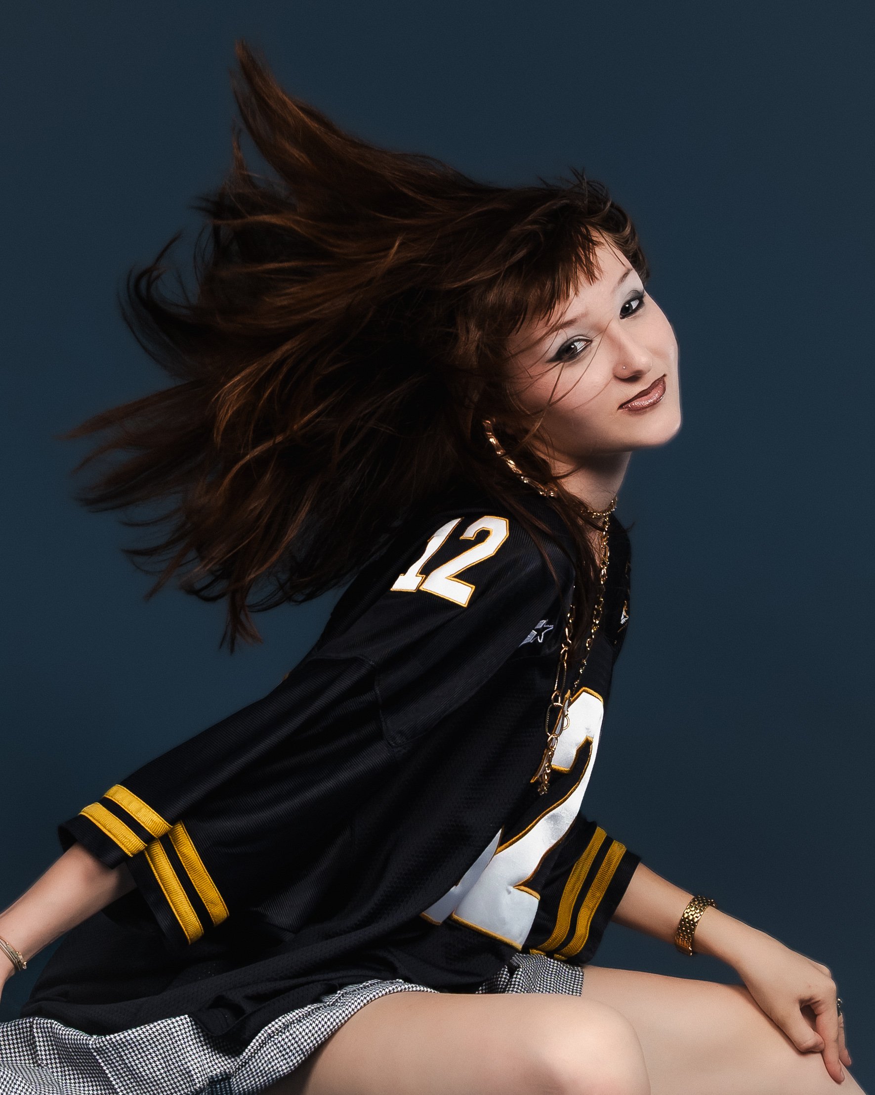 Une jeune femme avec des cheveux bruns longs en mouvement, portant un maillot de football américain noir avec le numéro 12, des accessoires dorés, assise contre un fond bleu foncé.