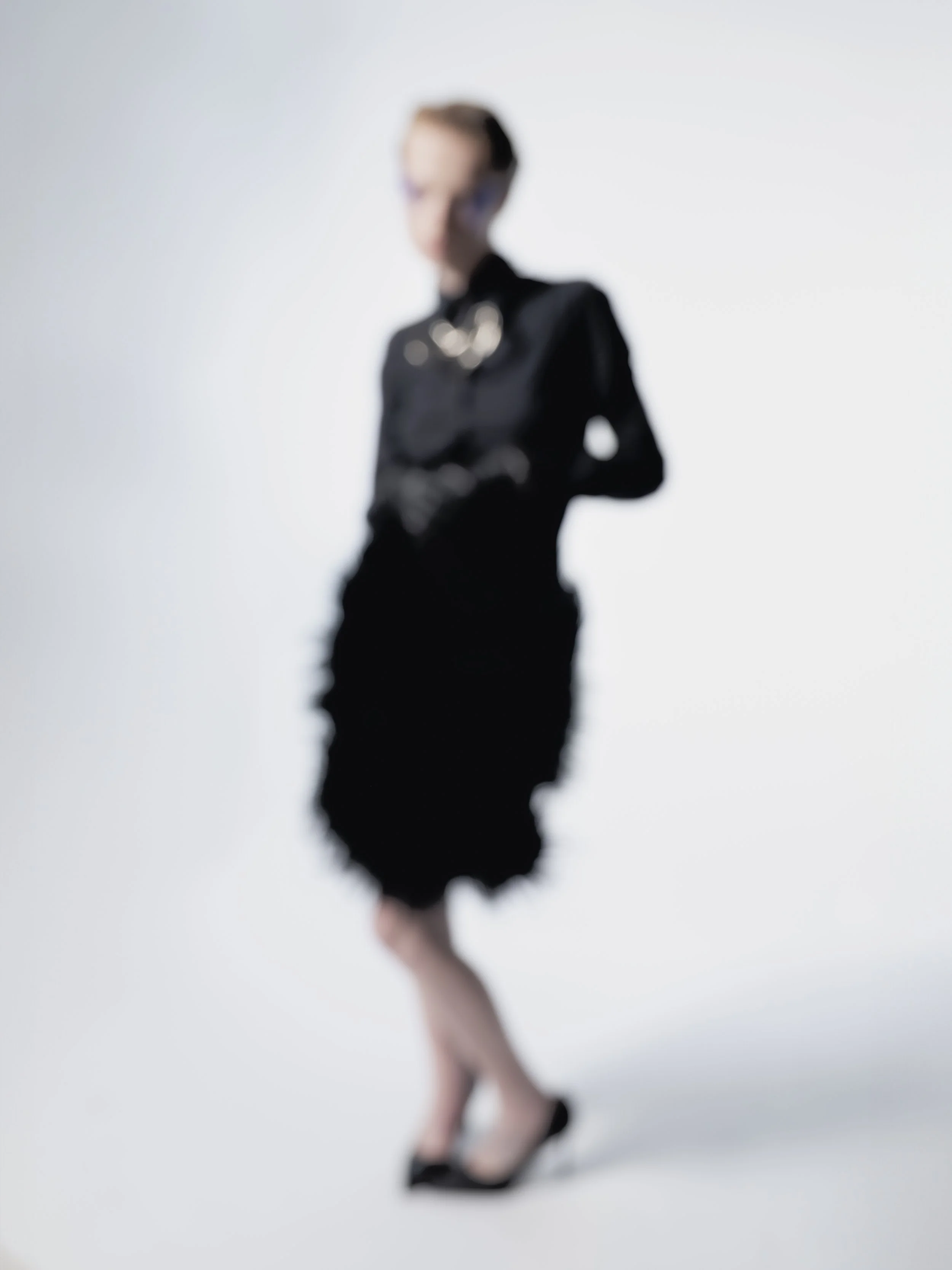 Une femme en robe noire, portant des talons, pose devant un fond clair.