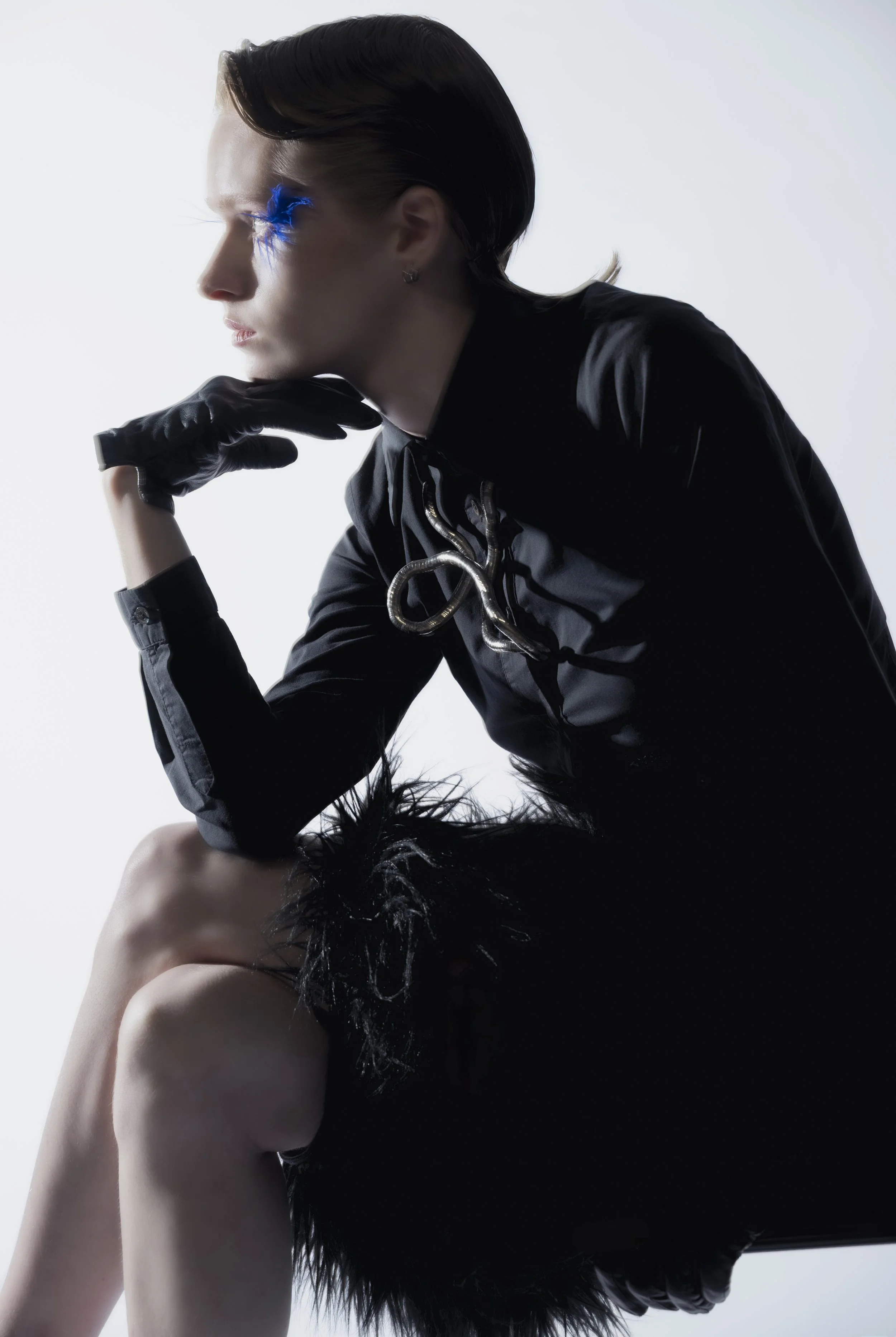 Une femme avec des cheveux noirs courts, portant une veste noire en satin et des gants en cuir noirs, pose en profil contre un fond blanc, avec un accessoire métallique autour du cou.