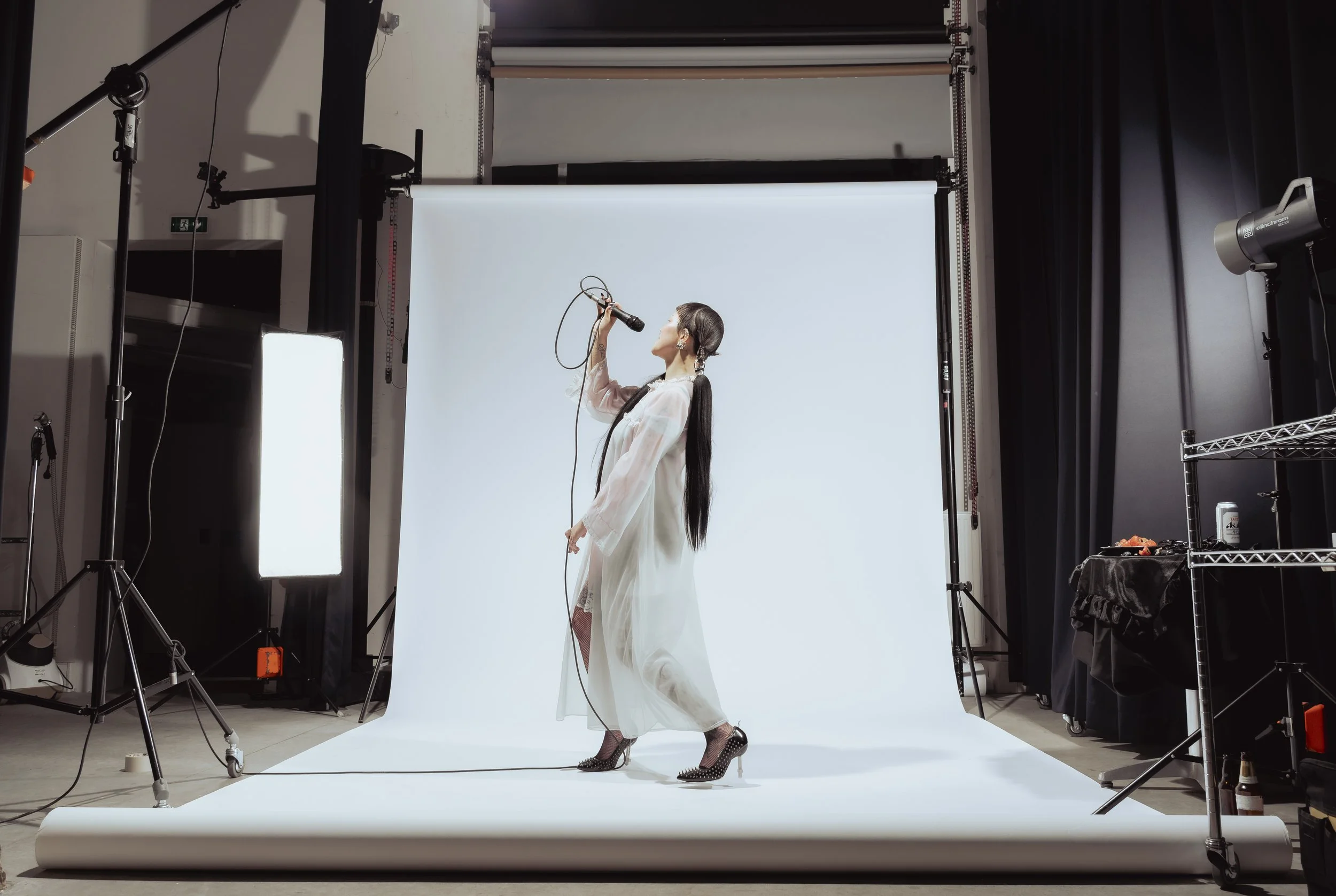 Une femme chante dans un studio photo avec un fond blanc, portant une robe longue et des talons à pointe avec des piquants.