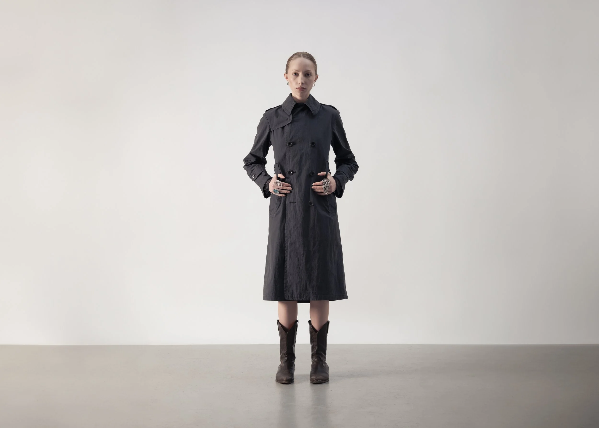 Femme portant un trench noir et des bottes, debout devant un fond gris clair.