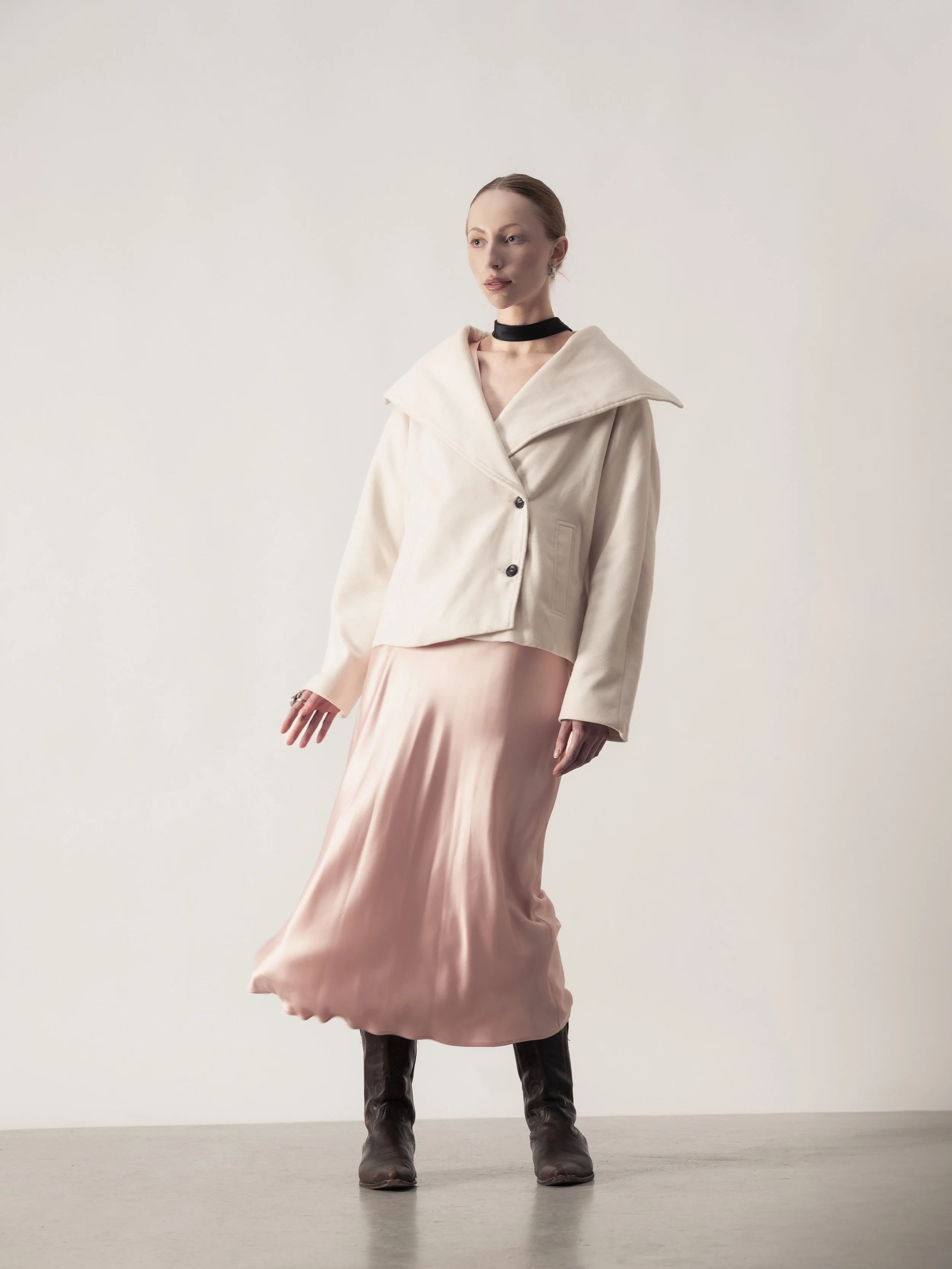 Une femme portant une veste beige, une jupe longue en satin rose pâle et des bottes en cuir noir, se tenant contre un fond neutre.