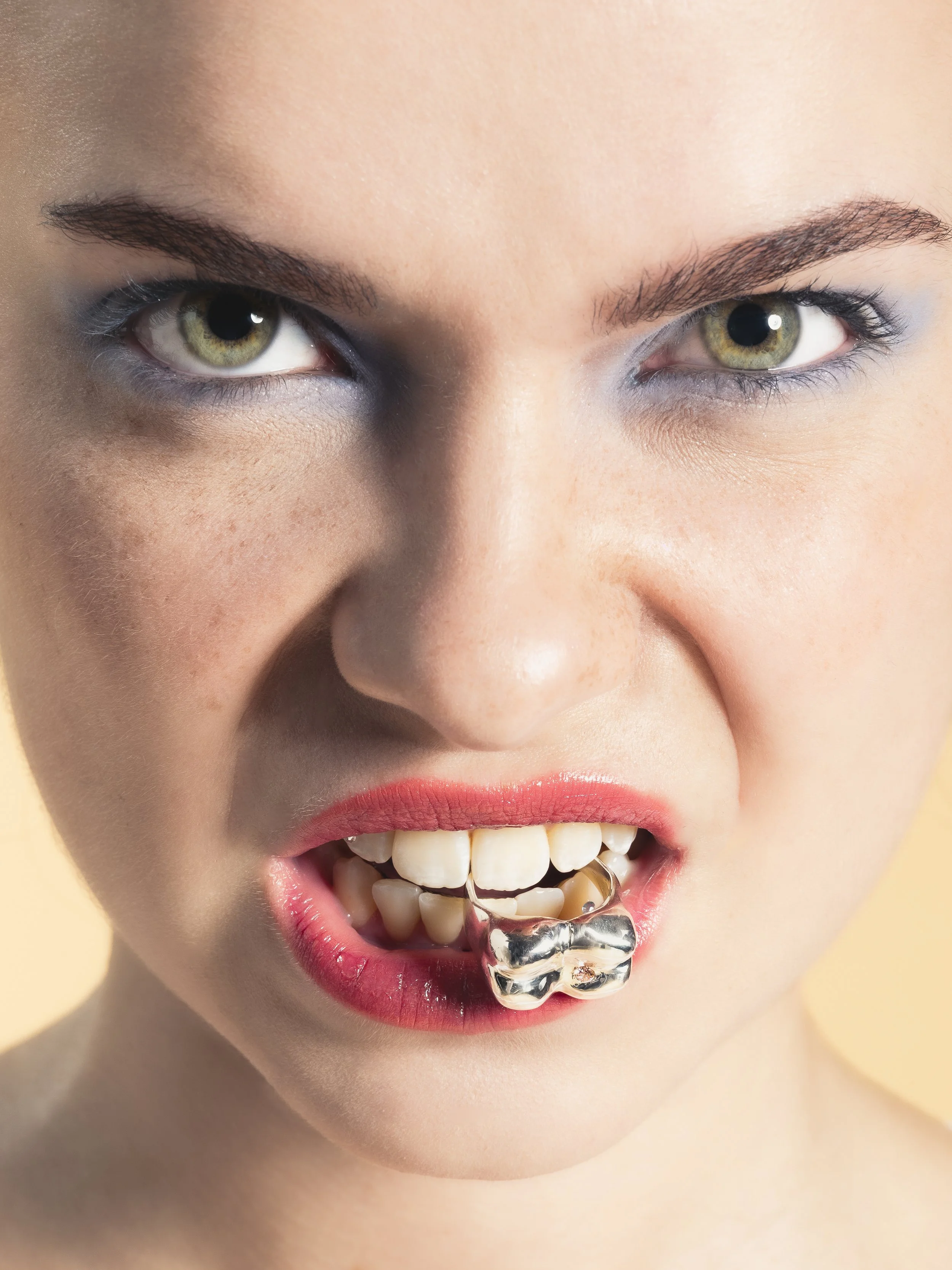 Gros plan d'une femme avec expression agressive, montrant ses dents, avec un bijou en forme d'os en or dans sa bouche.