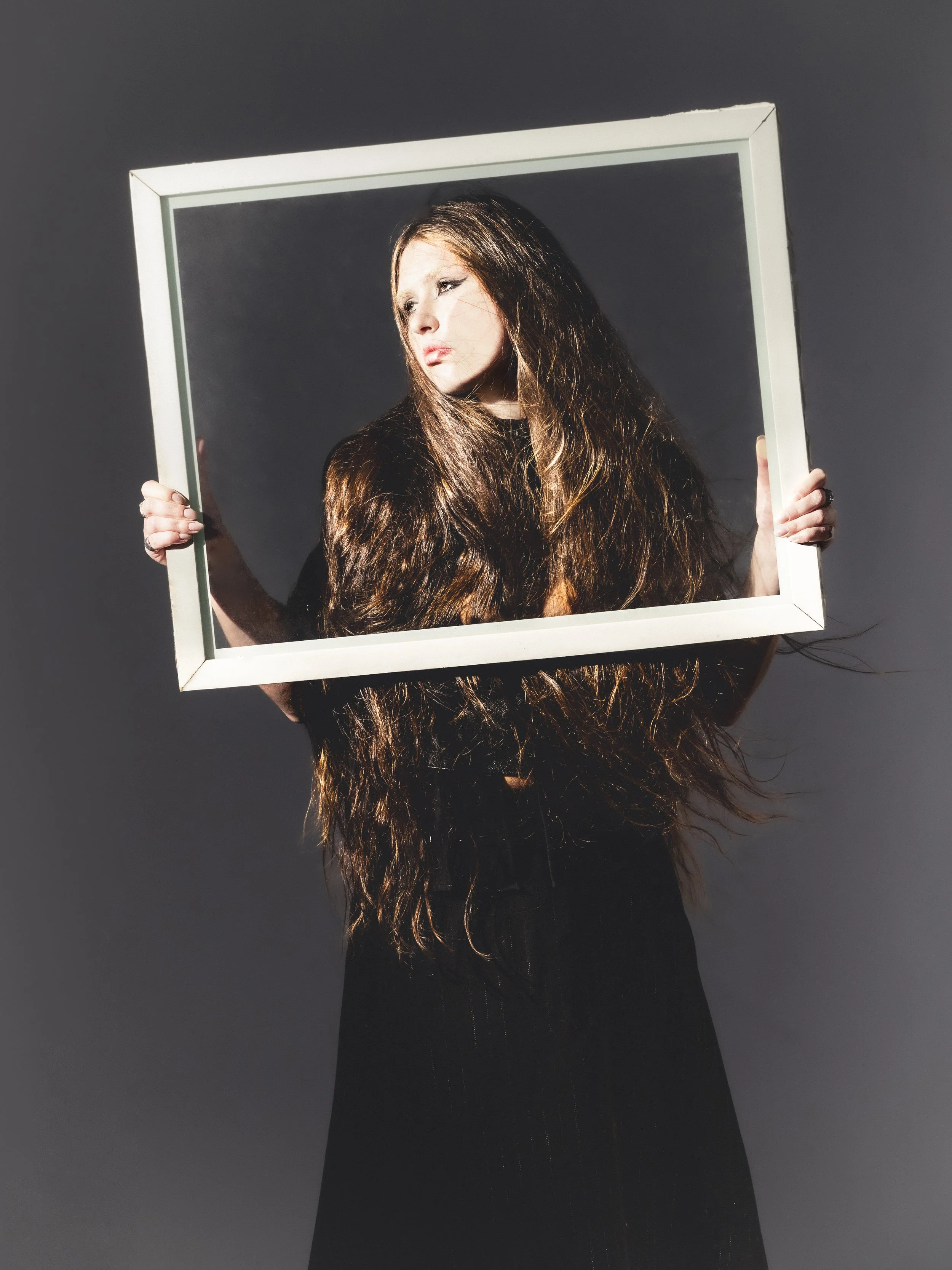 Une femme avec de longs cheveux bruns tient un cadre blanc devant elle, semblant regarder à travers. Elle porte des vêtements noirs, et le fond est gris.