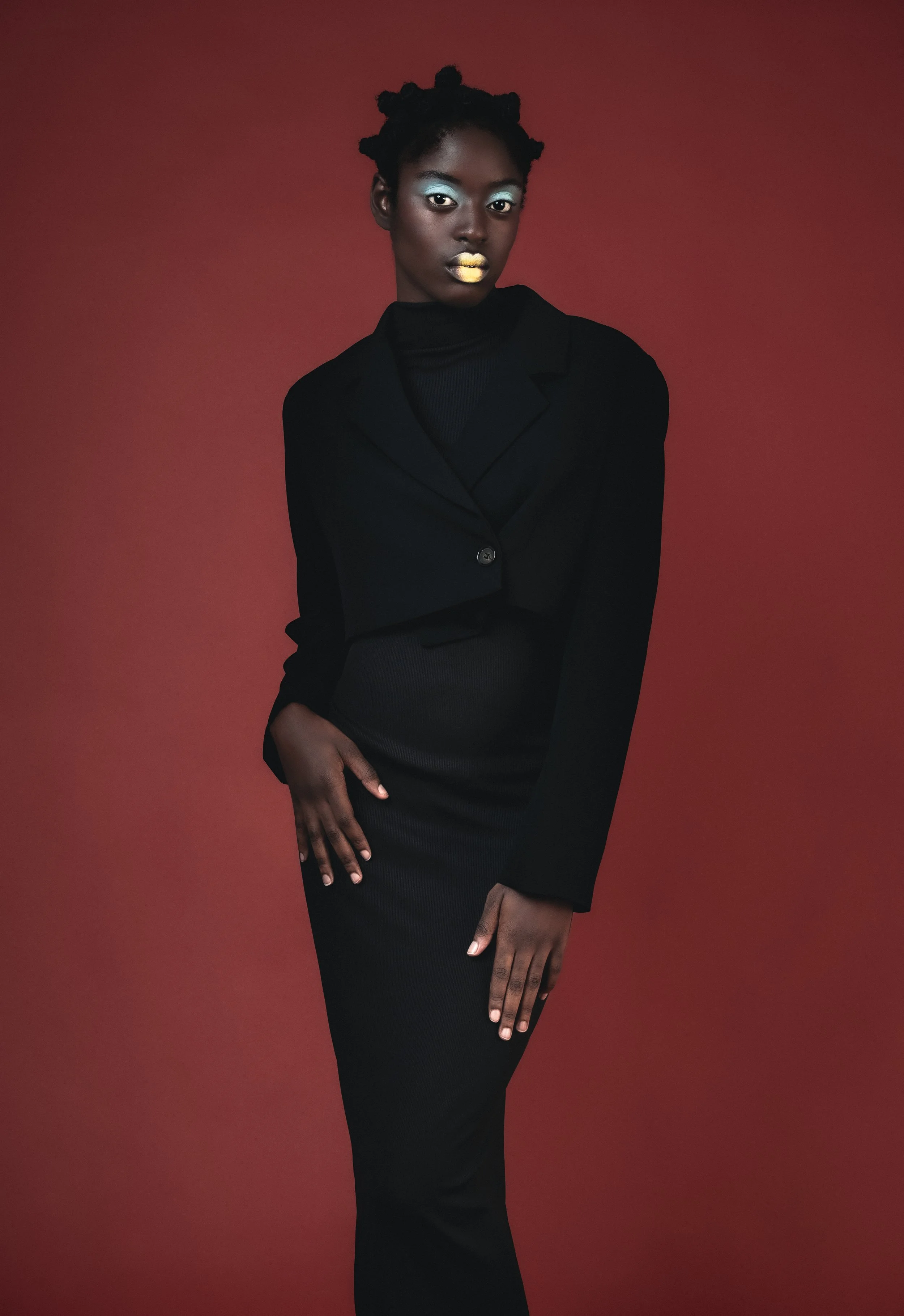 Une femme portant un tailleur noir avec un fond rouge. Elle a les cheveux en bantu knots et porte du maquillage avec des couleurs vives, notamment du bleu sur les paupières et du jaune sur les lèvres.