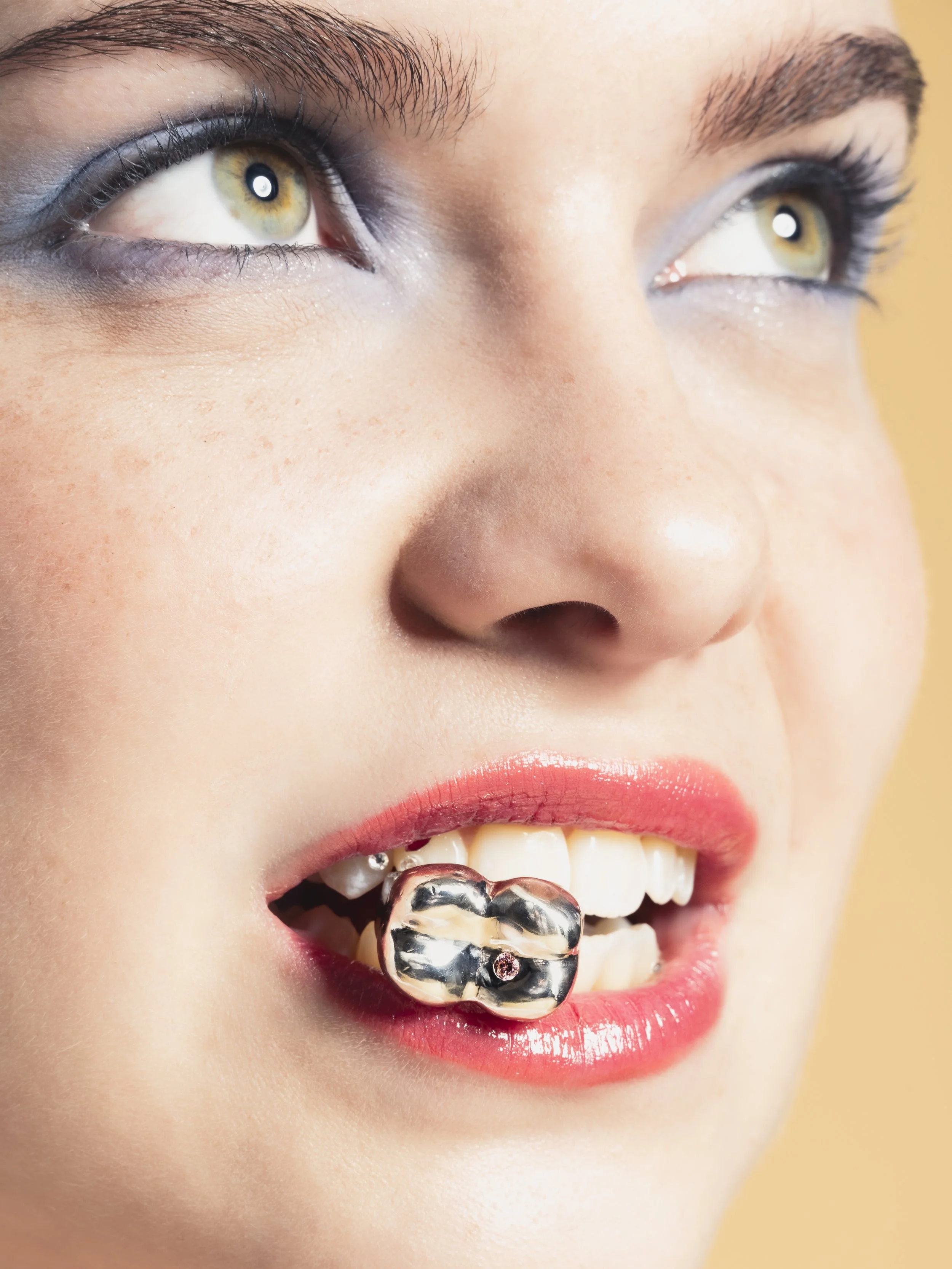 Gros plan d'une femme avec un maquillage coloré, portant un bijou en forme de dent sur ses dents.