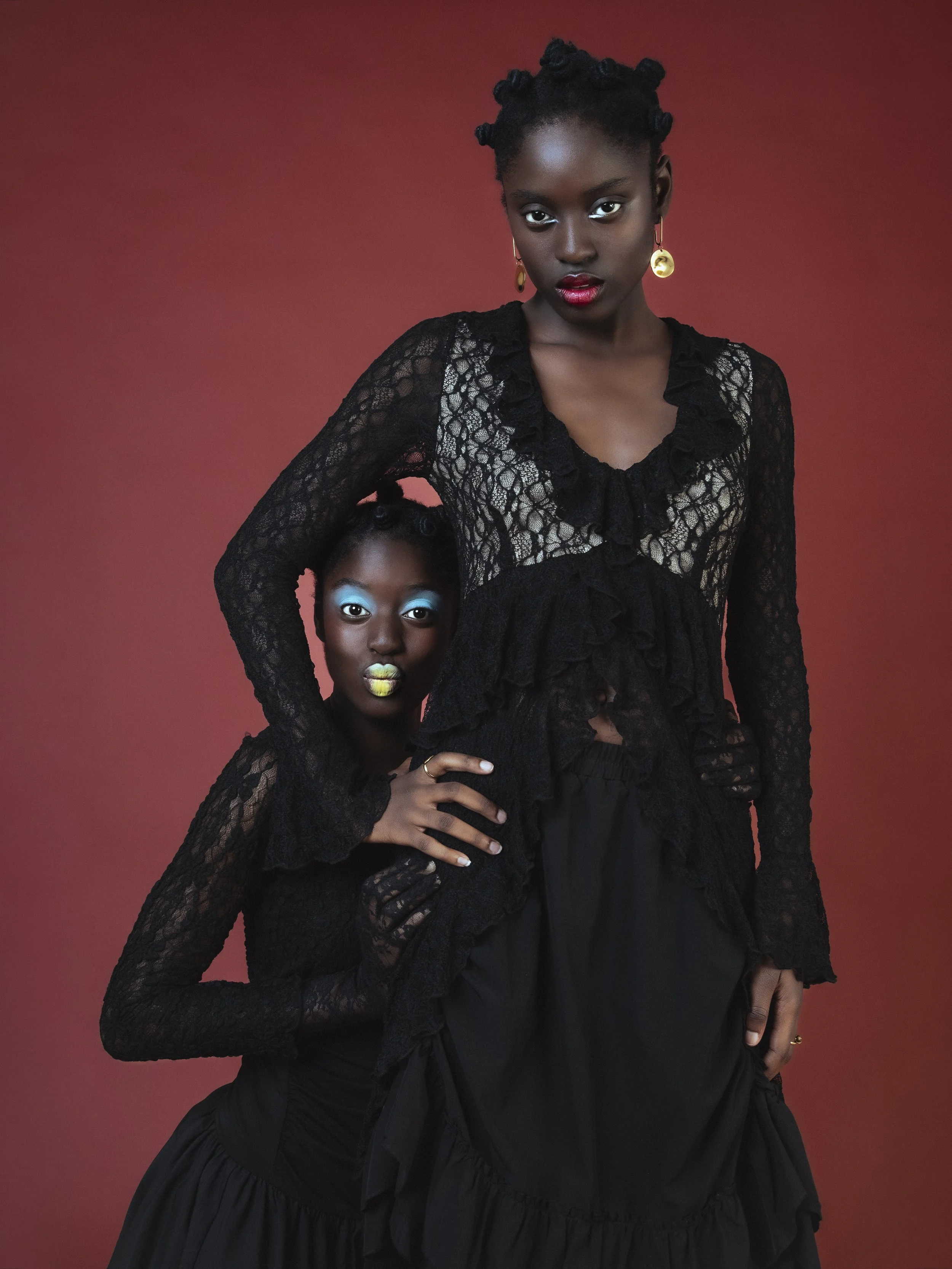 Deux jeunes femmes noires posant contre un fond rouge, portant des tenues en dentelle noire avec maquillage distinctif, l'une debout et l'autre à genoux, la plus petite tenant le nombril de l'autre.