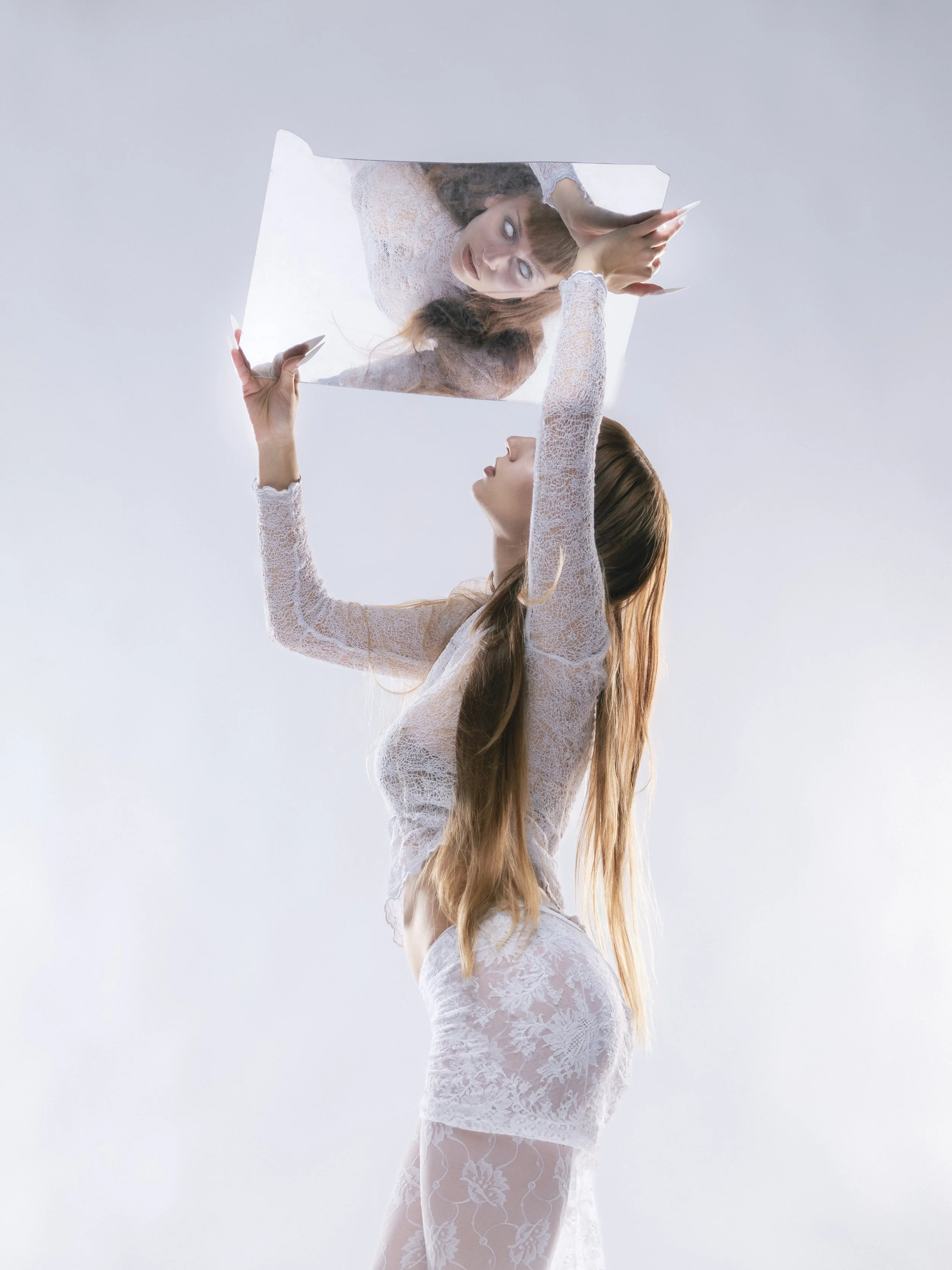Femme portant une robe en dentelle blanche tenant un miroir, reflétant son visage et ses bras dans le miroir.