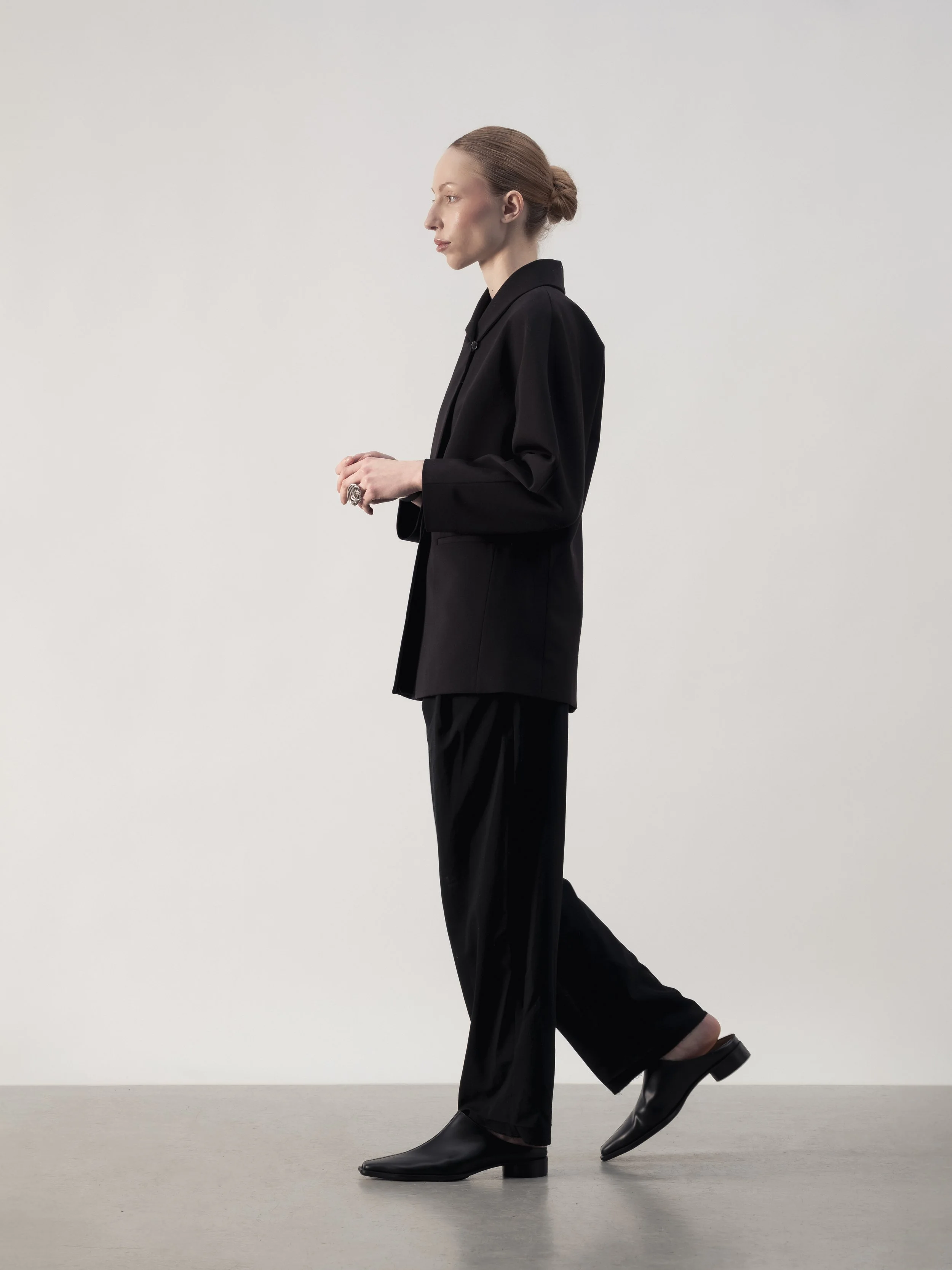 Femme en costume noir, debout sur un fond blanc, vue de profil.