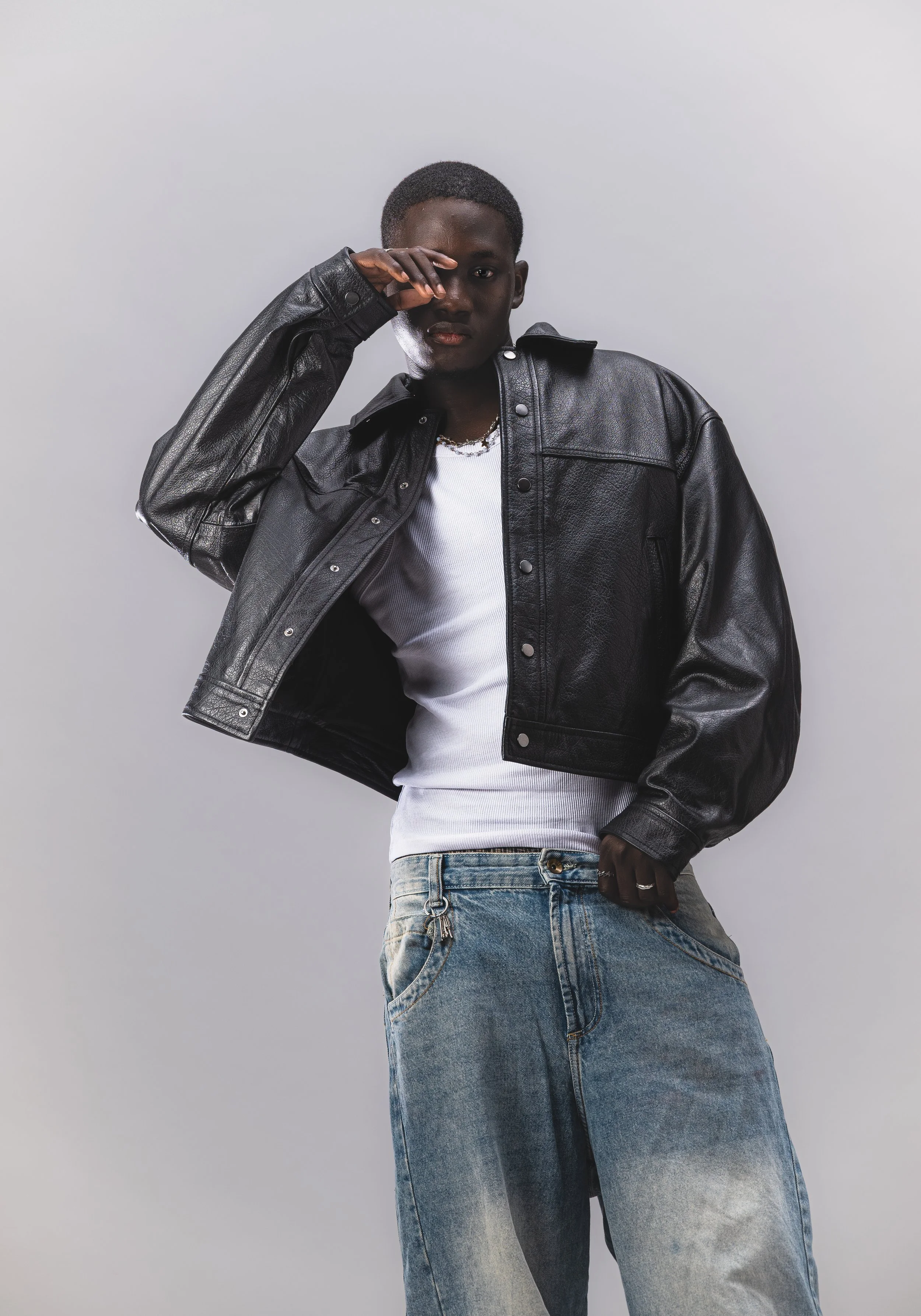 Jeune homme portant une veste en cuir noire, un t-shirt blanc et un jean délavé, posant avec une main près du visage contre un fond gris clair.