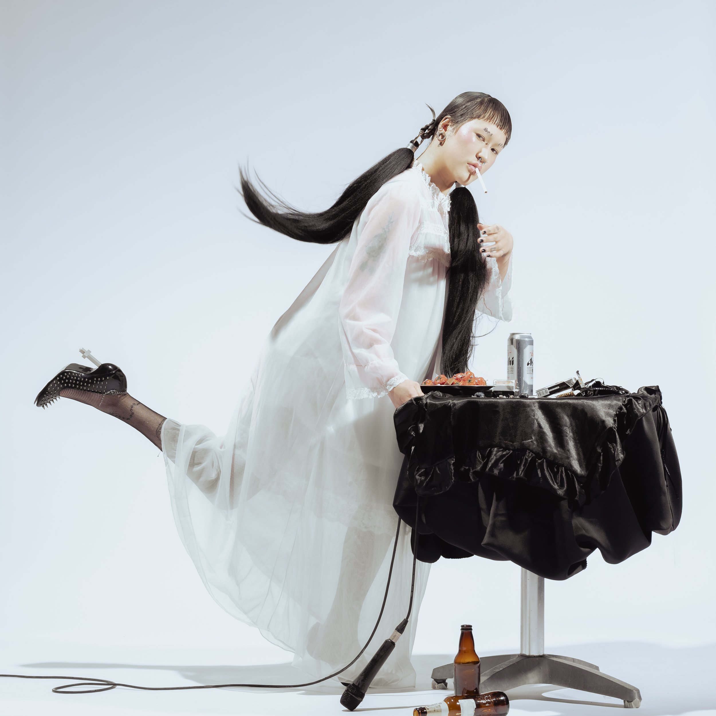 Une femme en robe blanche, avec de longues tresses noires, tient une cigarette et se trouve près d'une table recouverte d'un tissu noir, avec des boissons et des objets dessus. L'ambiance est neutre avec un fond blanc.