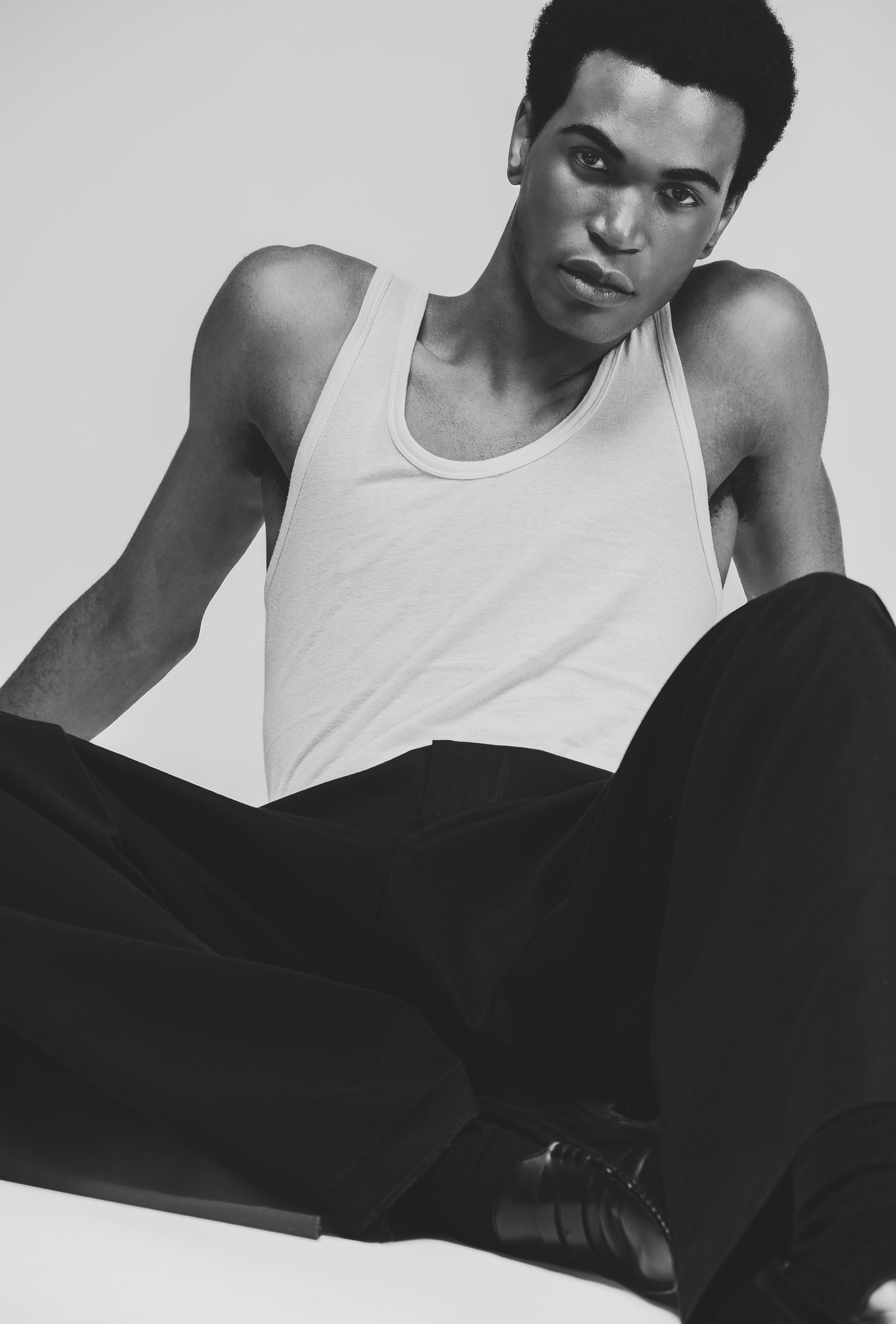 Portrait en noir et blanc d'une femme aux cheveux courts et bouclés, portant un débardeur blanc et un pantalon noir, assise dans une pose décontractée.