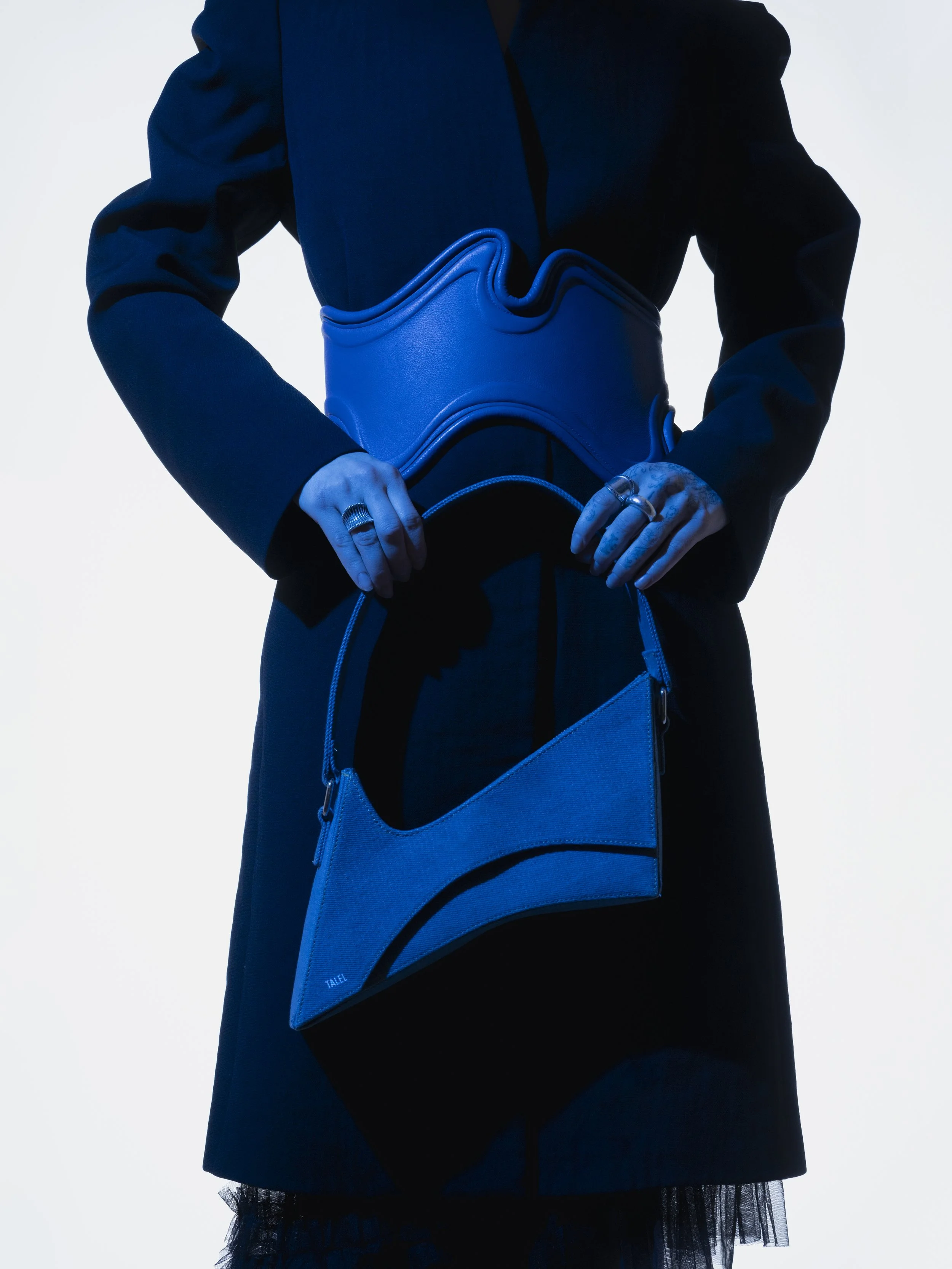 Une personne porte une robe longue noire avec une ceinture bleue et tient un sac à main bleu, sur fond blanc.